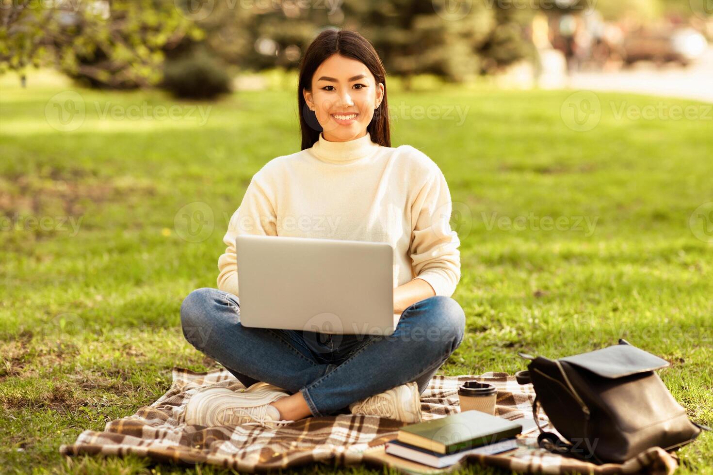 Asian woman is sitting on a colorful blanket outdoors, typing on a laptop computer. She appears focused and engaged in her work, with trees and grass in the background photo