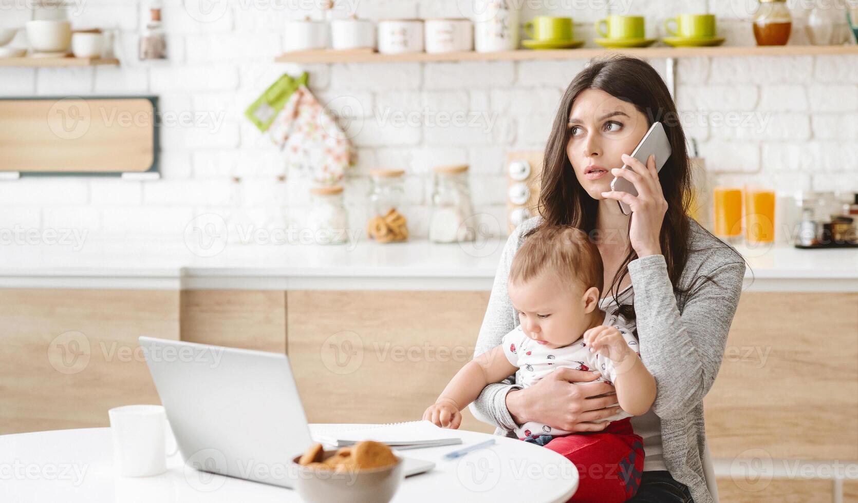 A woman sits in her kitchen with a baby in her arms. She is on the phone, looking concerned, with her laptop open in front of her. The kitchen is bright and modern photo