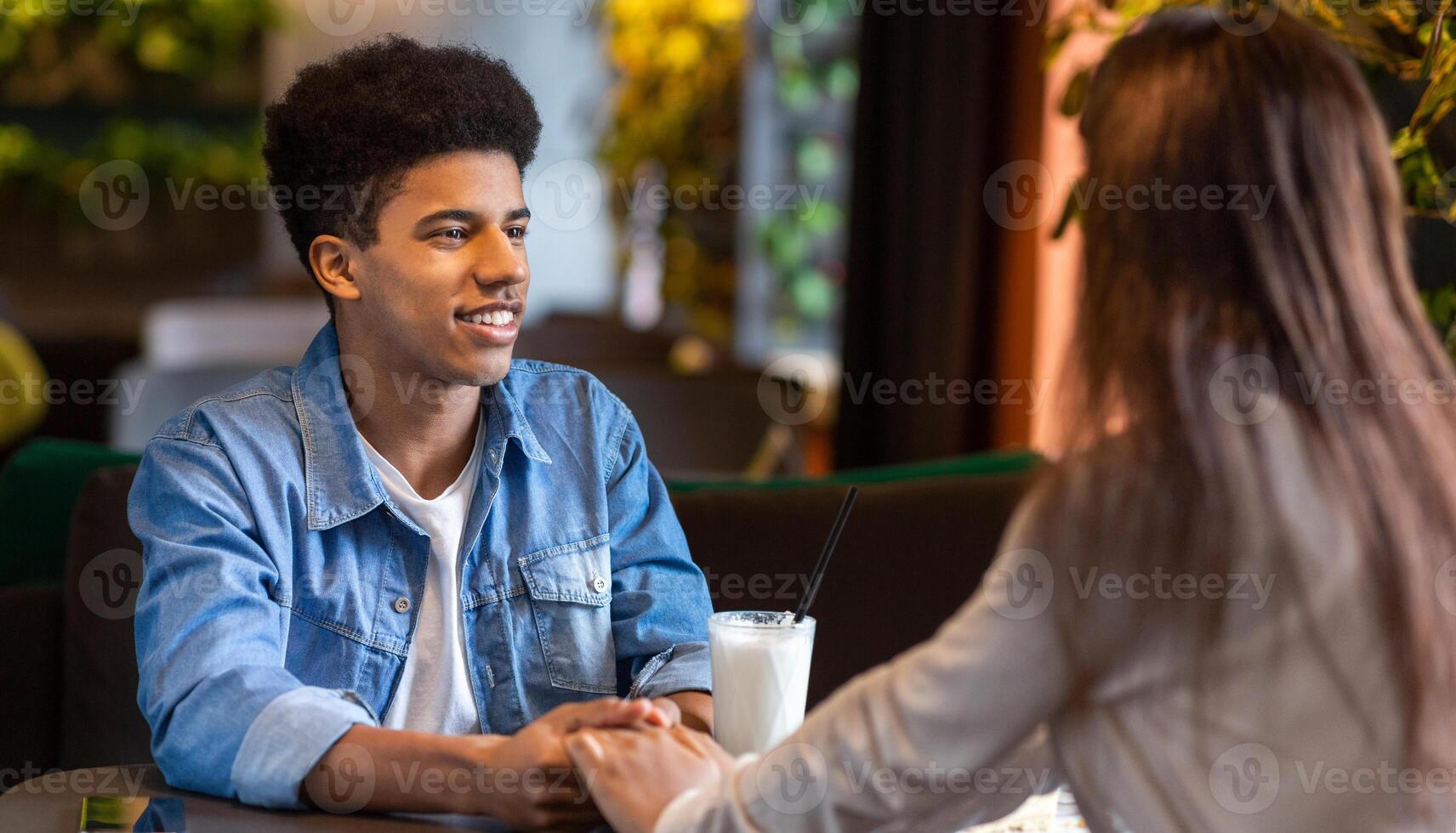 Black guy and girl are engaged in a conversation at a table in a restaurant. The guy appears to be speaking while the girl listens attentively. The setting suggests a casual dining environment. photo