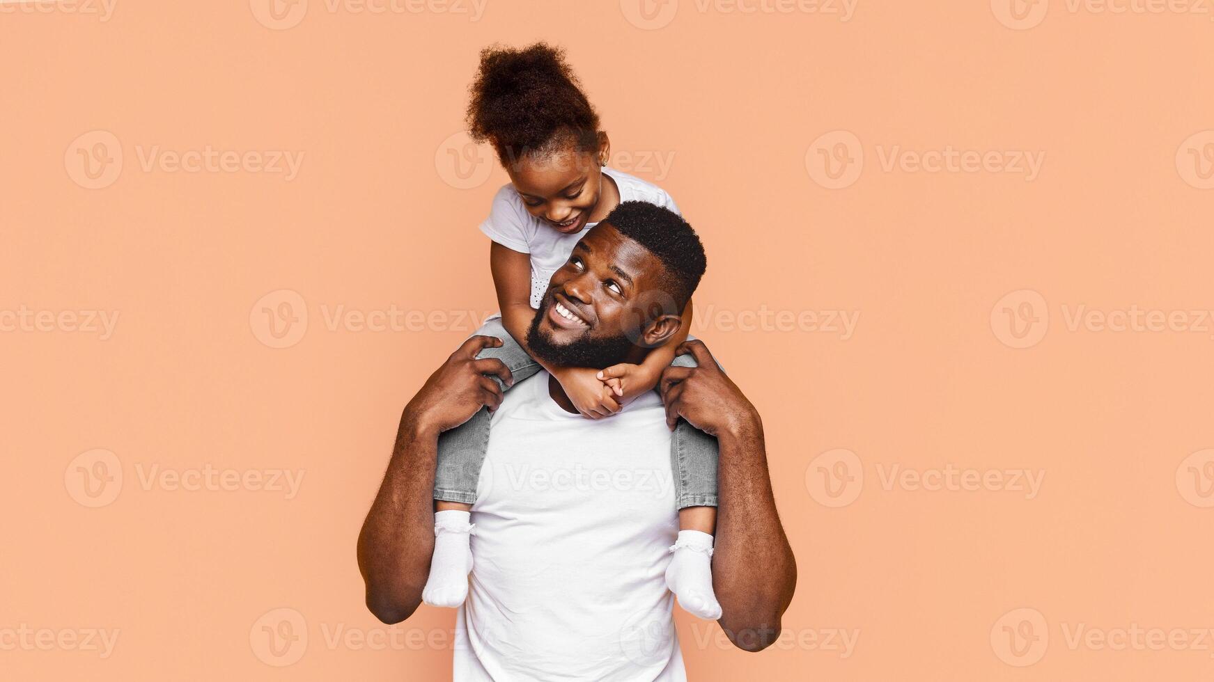 Happy African American father and his daughter posing together against a peach background. The father is smiling and looking to the side while holding his daughter on his shoulders photo