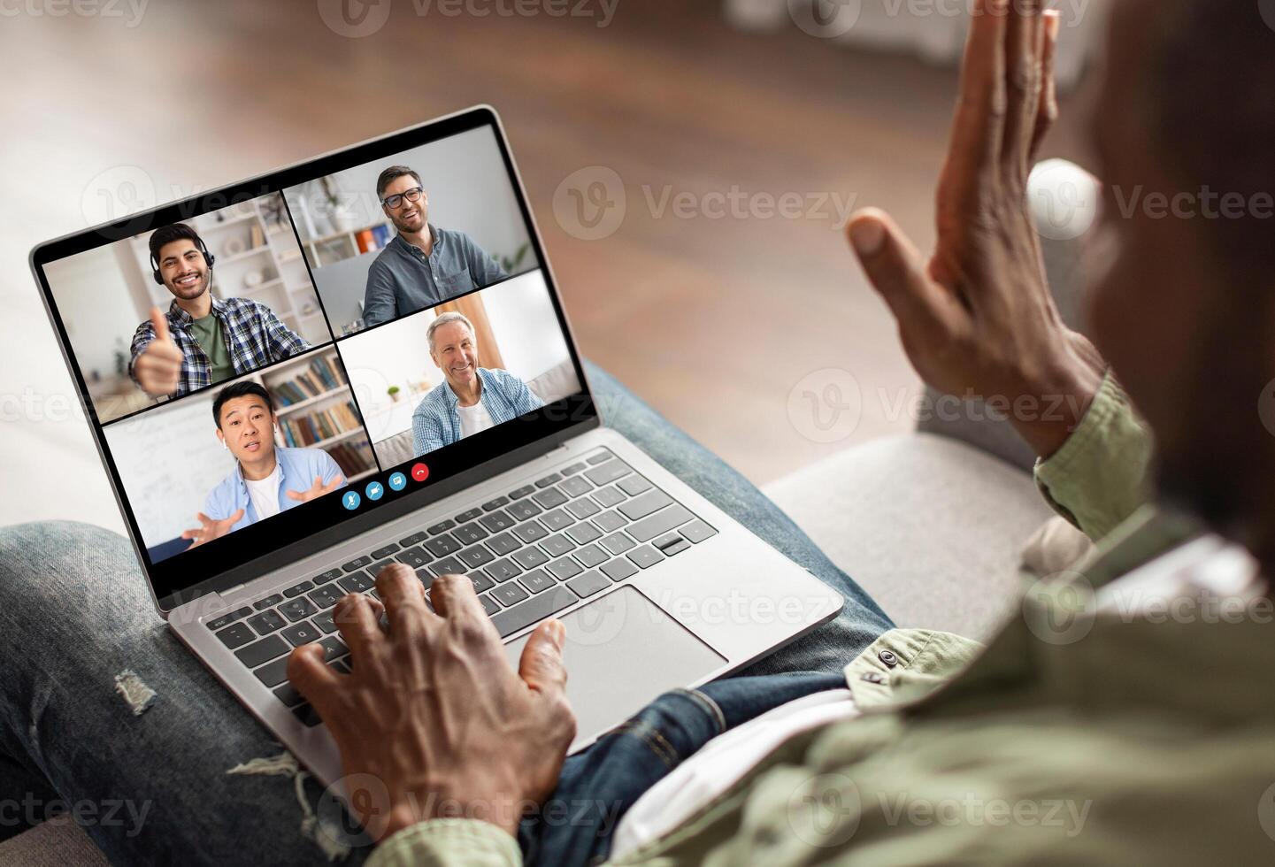 A man is sitting on a couch and participating in a conference call with three other colleagues on his laptop. He is using his hand to gesture and is clearly engaged in the conversation photo