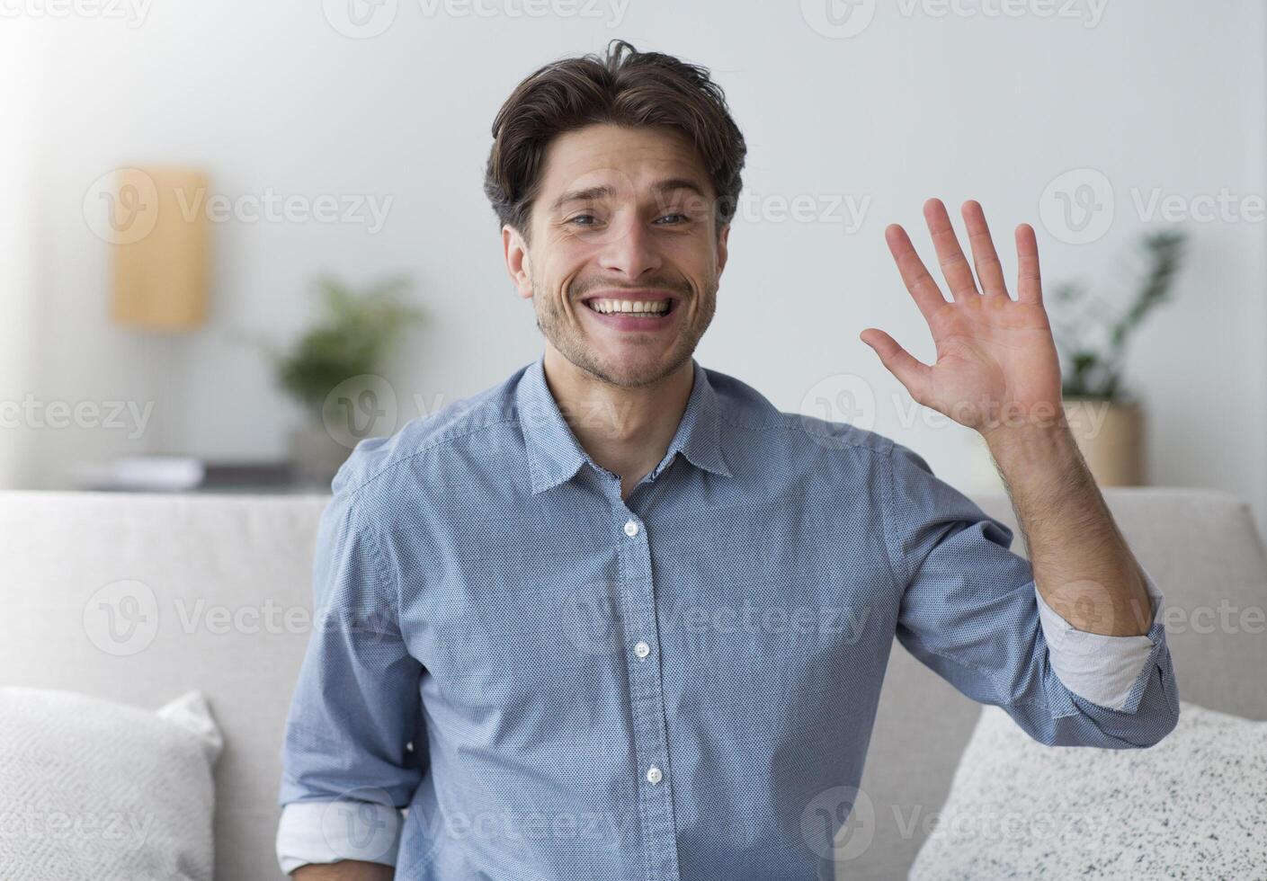 Hello. Smiling Man Waving Hand Looking At Camera Sitting On Sofa Indoor. photo