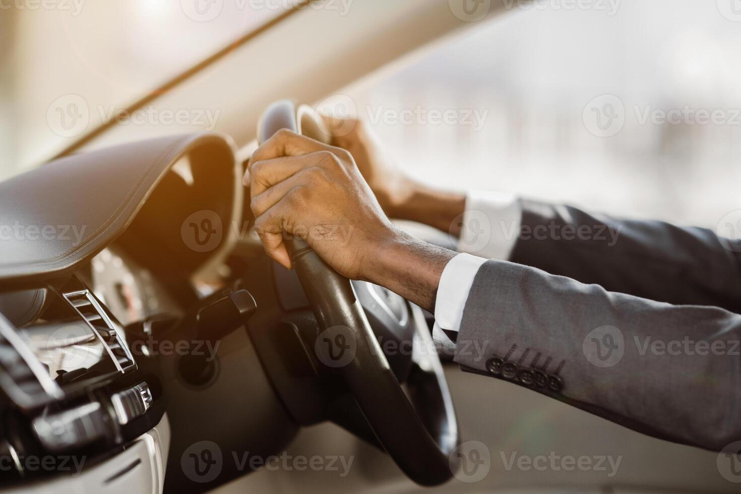 This image shows a closeup of a persons hands gripping the steering wheel of a car. The driver is wearing a suit jacket and their hands are visible in the foreground photo