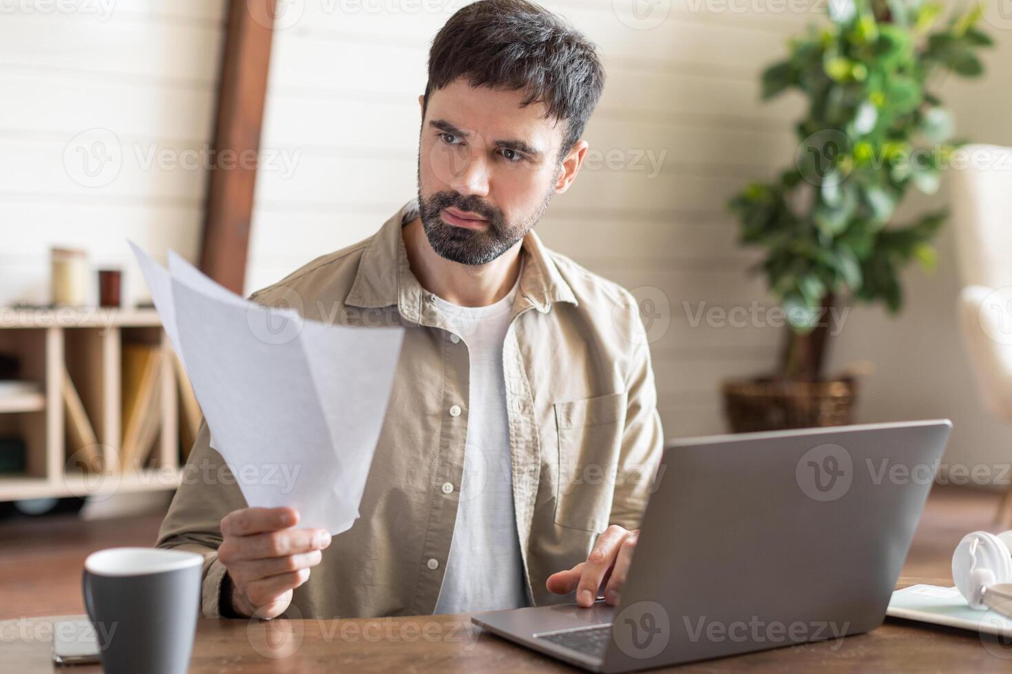 A man is seated at a table, focused on his laptop screen, surrounded by scattered papers. He appears engaged in work or study, possibly in a home or office setting. photo
