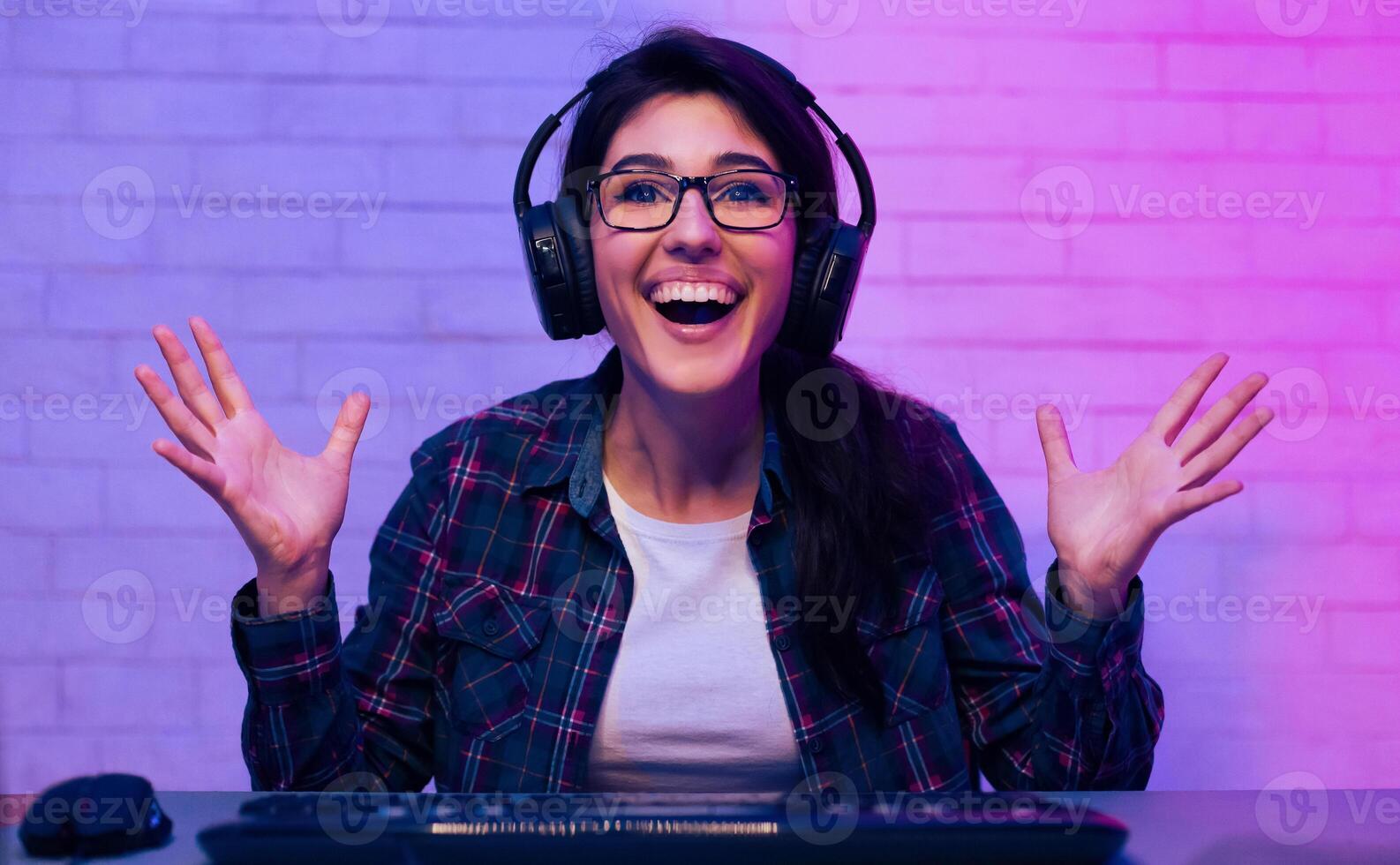 This is a photo of a woman wearing headphones and a plaid shirt, seated in front of a computer with her arms raised. She is smiling broadly and looks excited. She is likely playing a game.