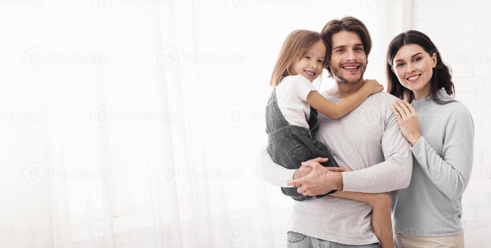 Family Insurance Concept. Portrait Of Happy Millennial Family Of Three With Little Daughter On Hands Posing Against Window At Home, Copy Space photo
