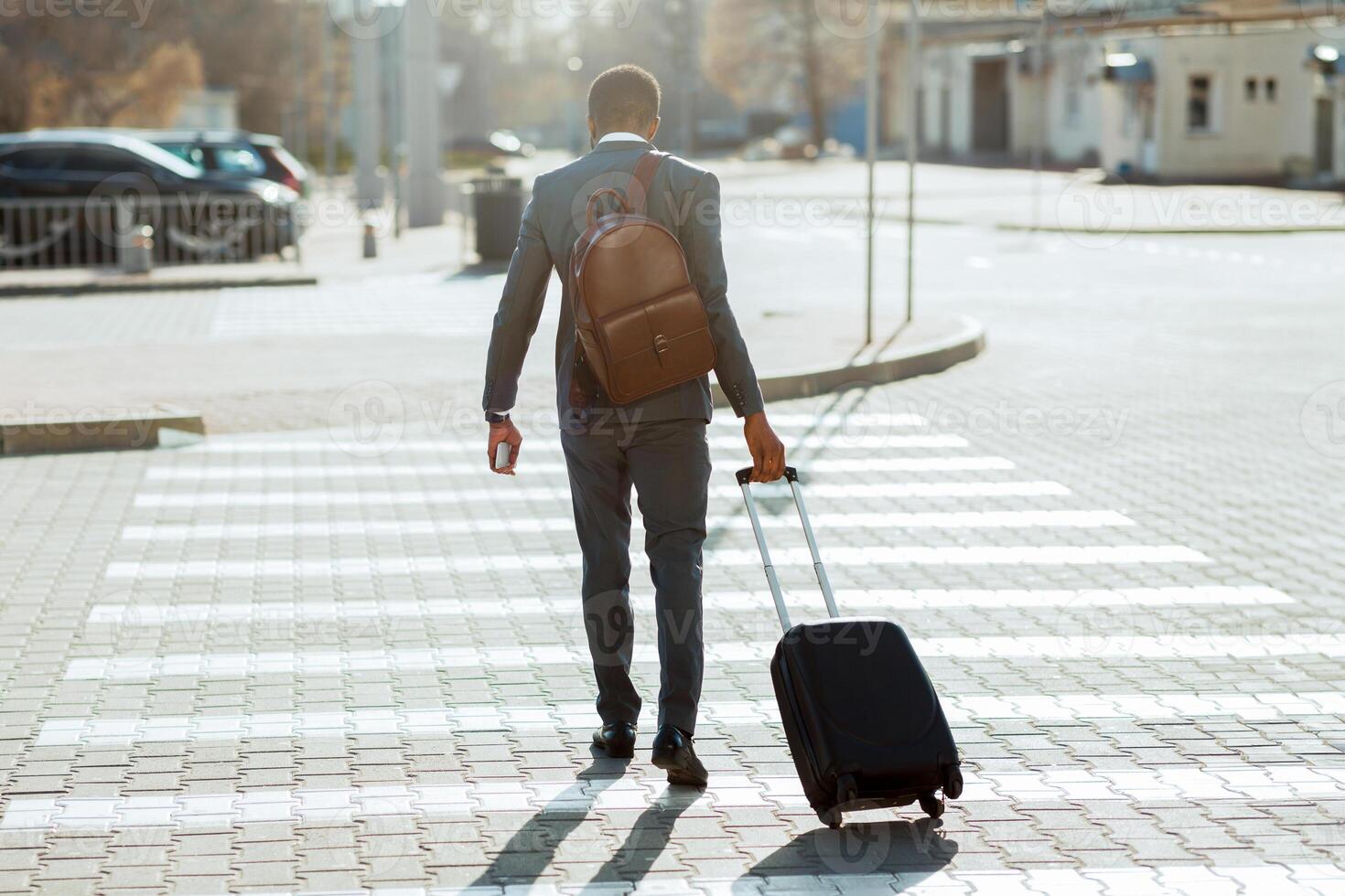 Black businessman in a suit crossing the street with a rolling suitcase, seen from behind in an urban setting. photo