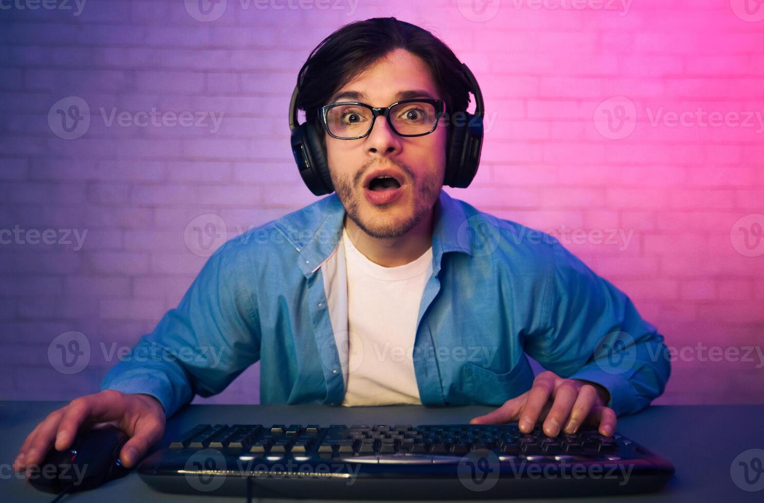 A young man wearing headphones and glasses sits in front of a keyboard and mouse, looking surprised and shocked. He appears to be playing a game at night in a dimly lit room. photo