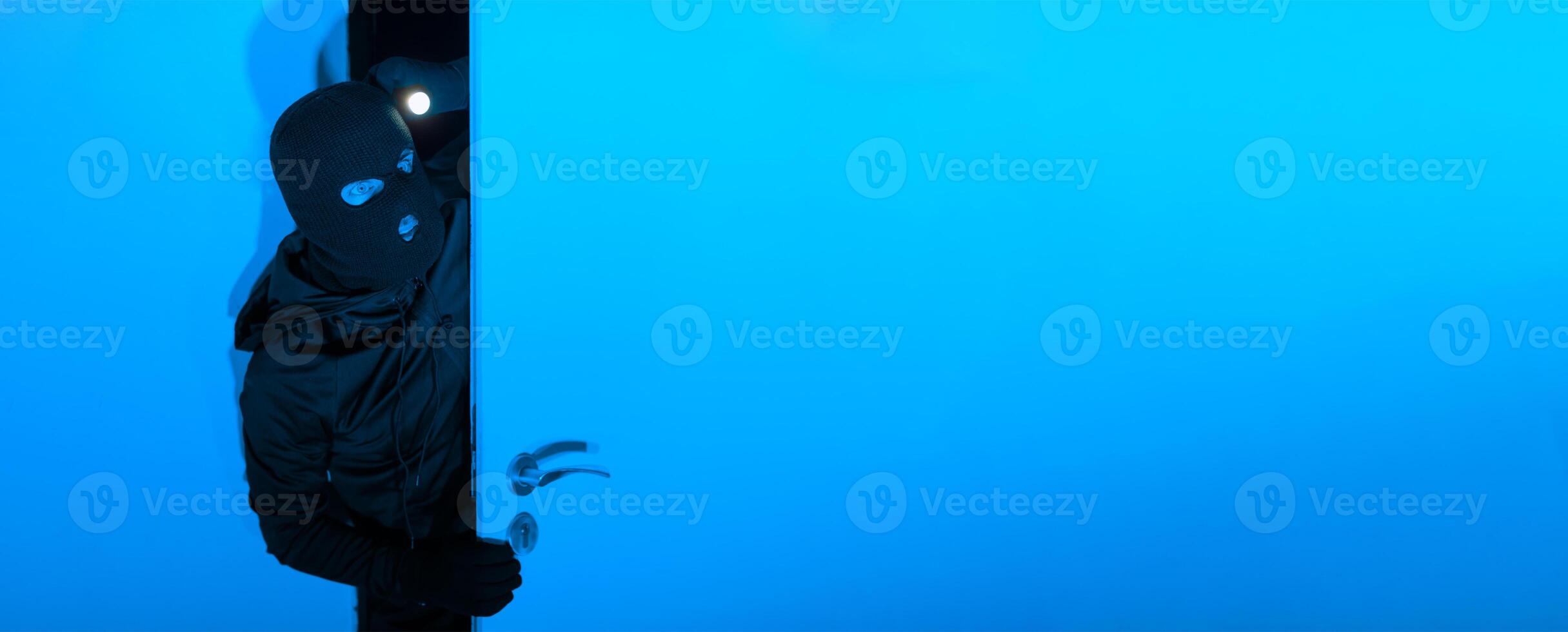 A cloaked individual cautiously opens a blue door in a darkened environment, illuminating the space with a flashlight, creating a suspenseful atmosphere of intrigue. photo