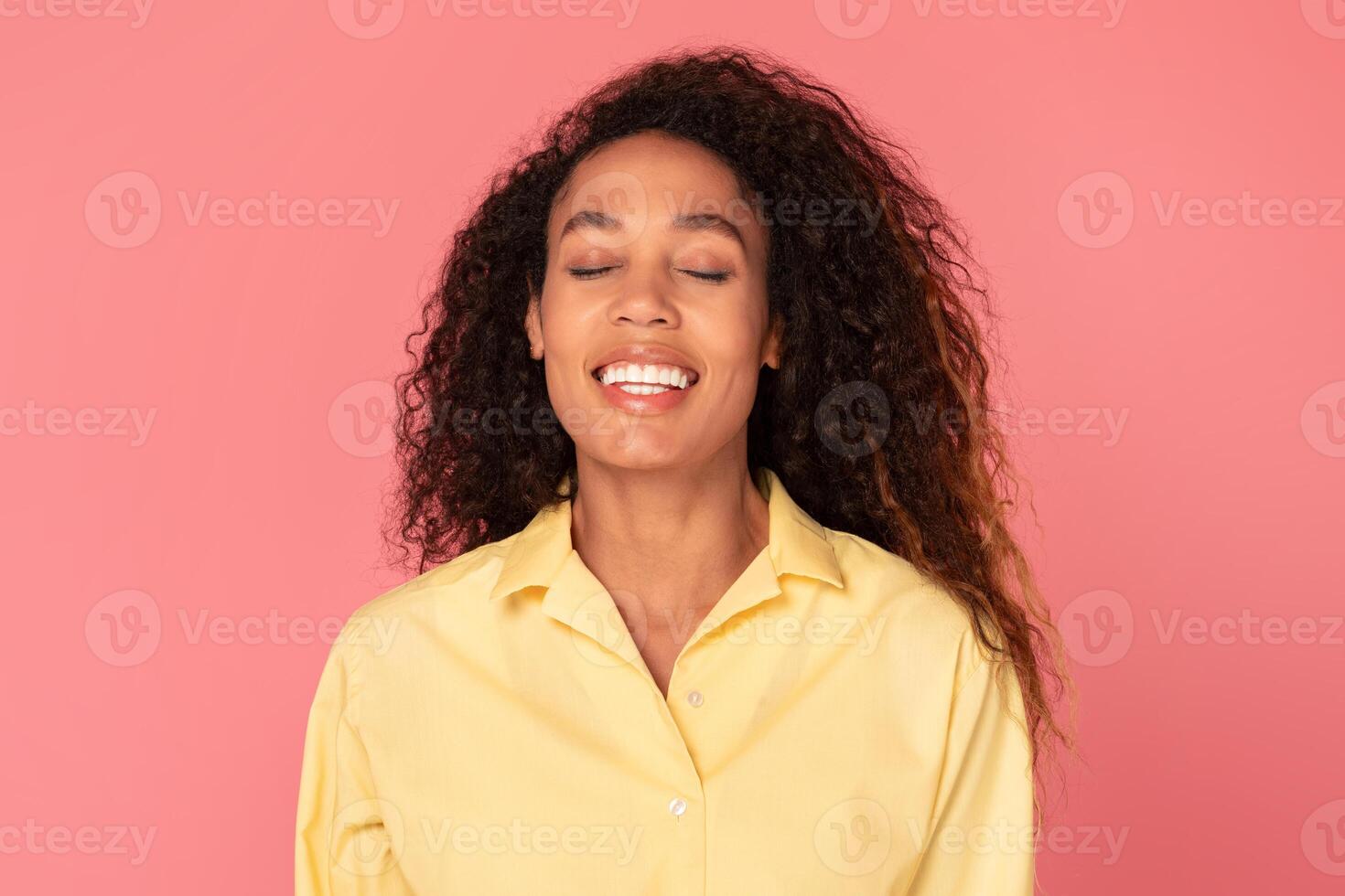 African American young woman with curly brown hair smiles broadly with her eyes closed while standing against a pink background. She is wearing a yellow button-down shirt. photo