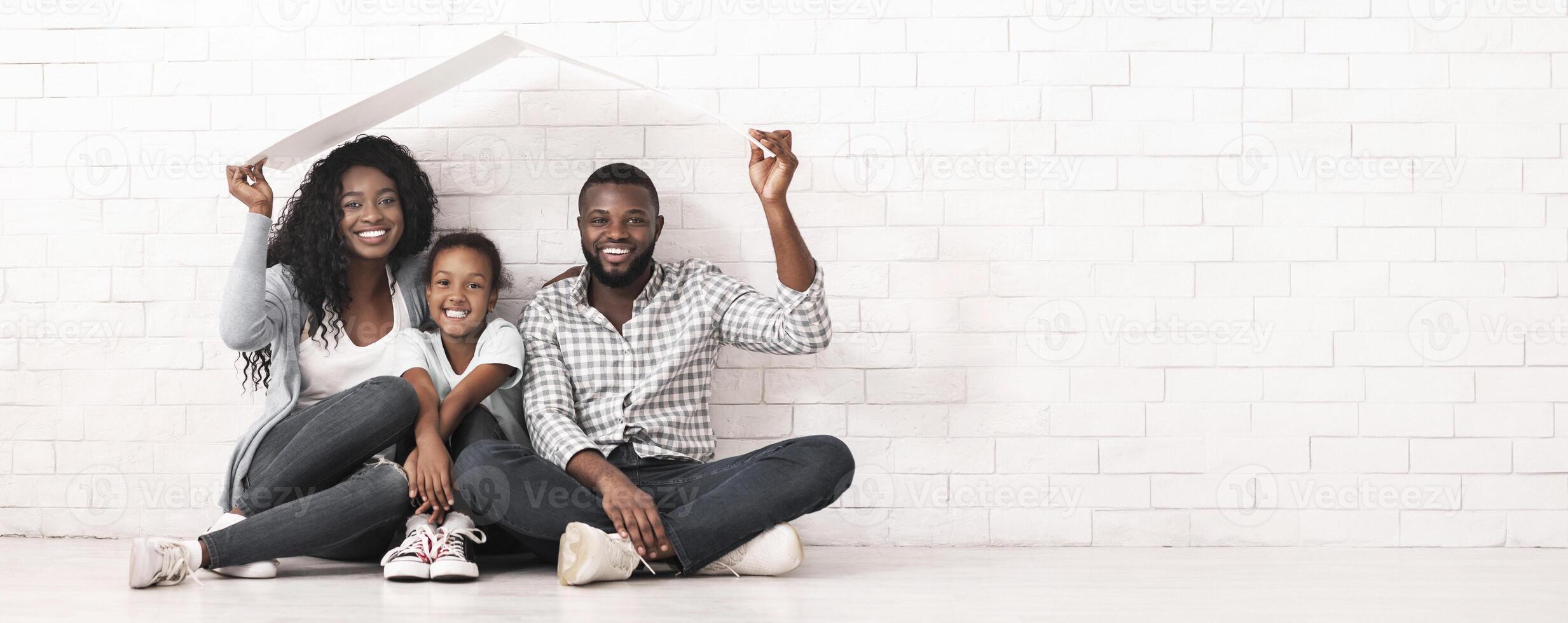 Family Housing Concept. Young Parents And Daughter Sitting Under Roof Dreaming Of New Home. White Brick Wall Background, Copy Space photo