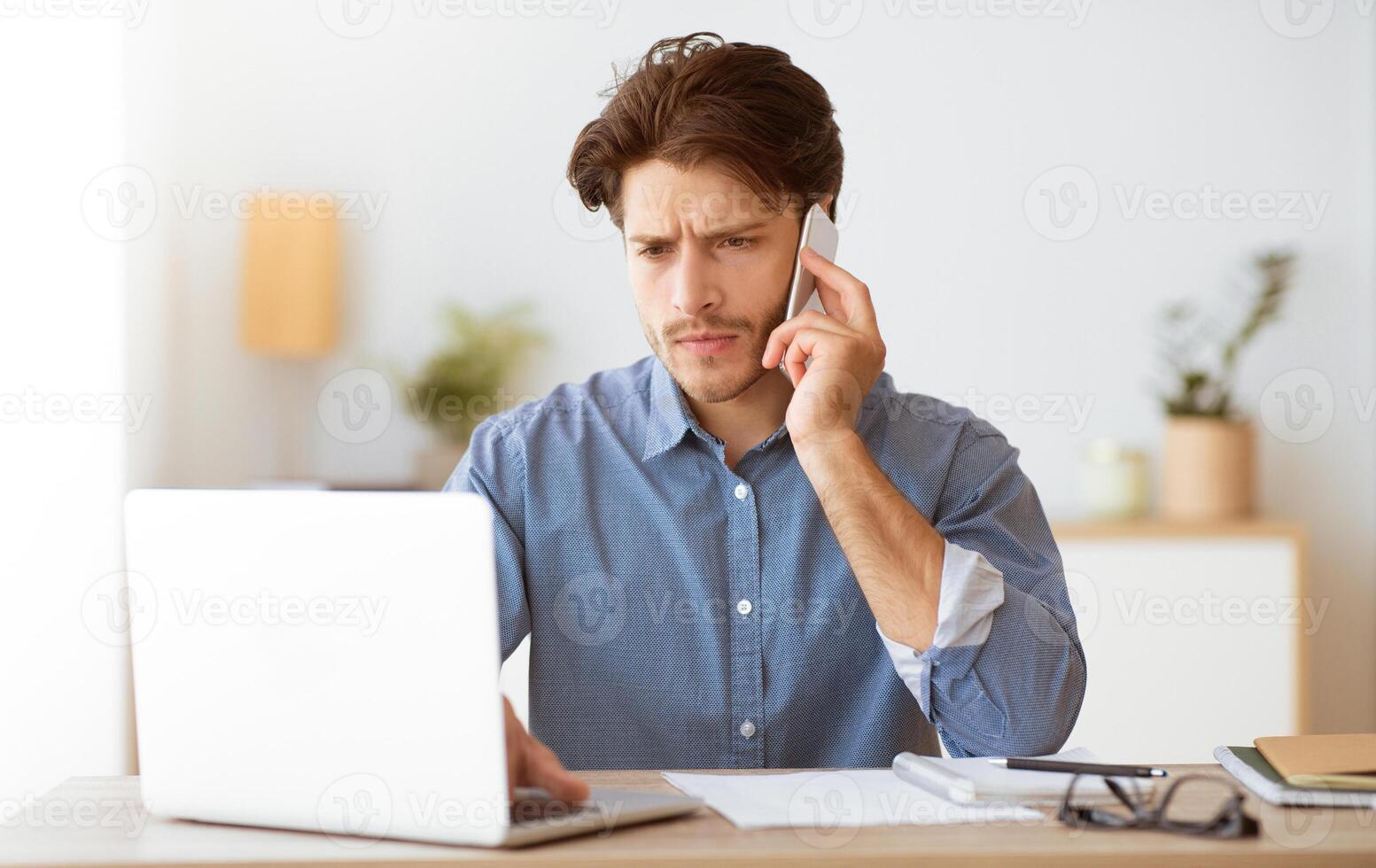 A man sits at a desk talking on his phone while working on his laptop. He looks focused and serious. photo