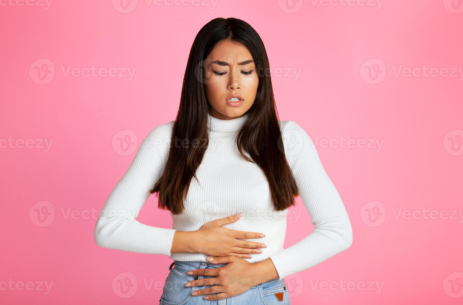 Asian young woman standing in front of a pink background. She is holding her stomach with both hands, and her expression is one of pain. She is likely experiencing a stomach ache or other discomfort. photo