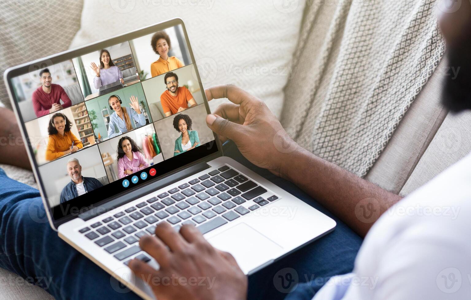 Black man sits on a couch, using a laptop to participate in a conference call with multiple other people. The man hands are visible as he engage with the screen. photo