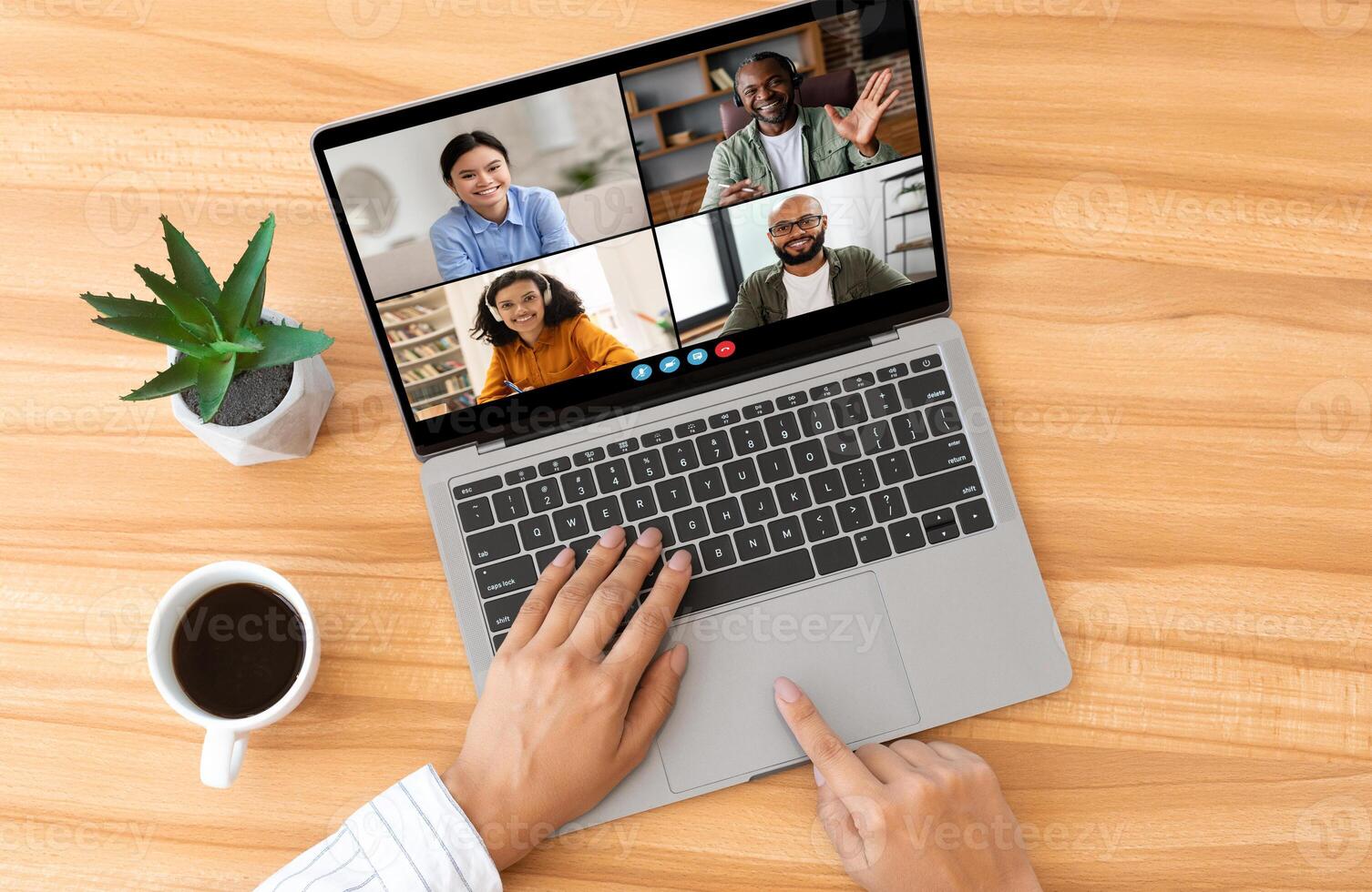 Woman using laptop for a chat with multiple participants. The laptop is open on a wooden desk with a cup of coffee and a potted plant nearby, top view photo