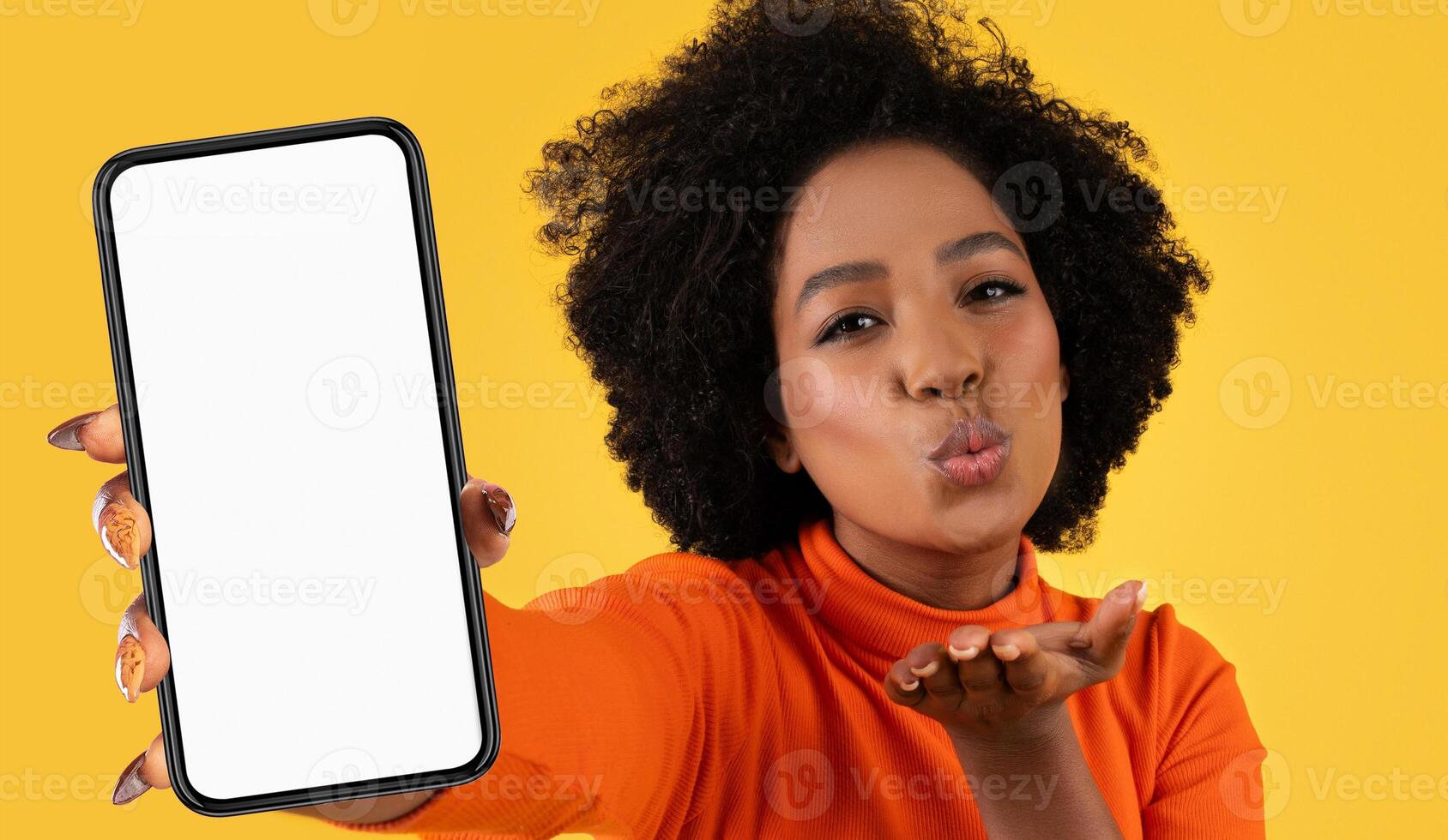 A young Black woman holding a smartphone in front of her and blowing a kiss. Her right hand is holding the phone, while her left hand is shaped into a kiss and pointed towards the viewer photo