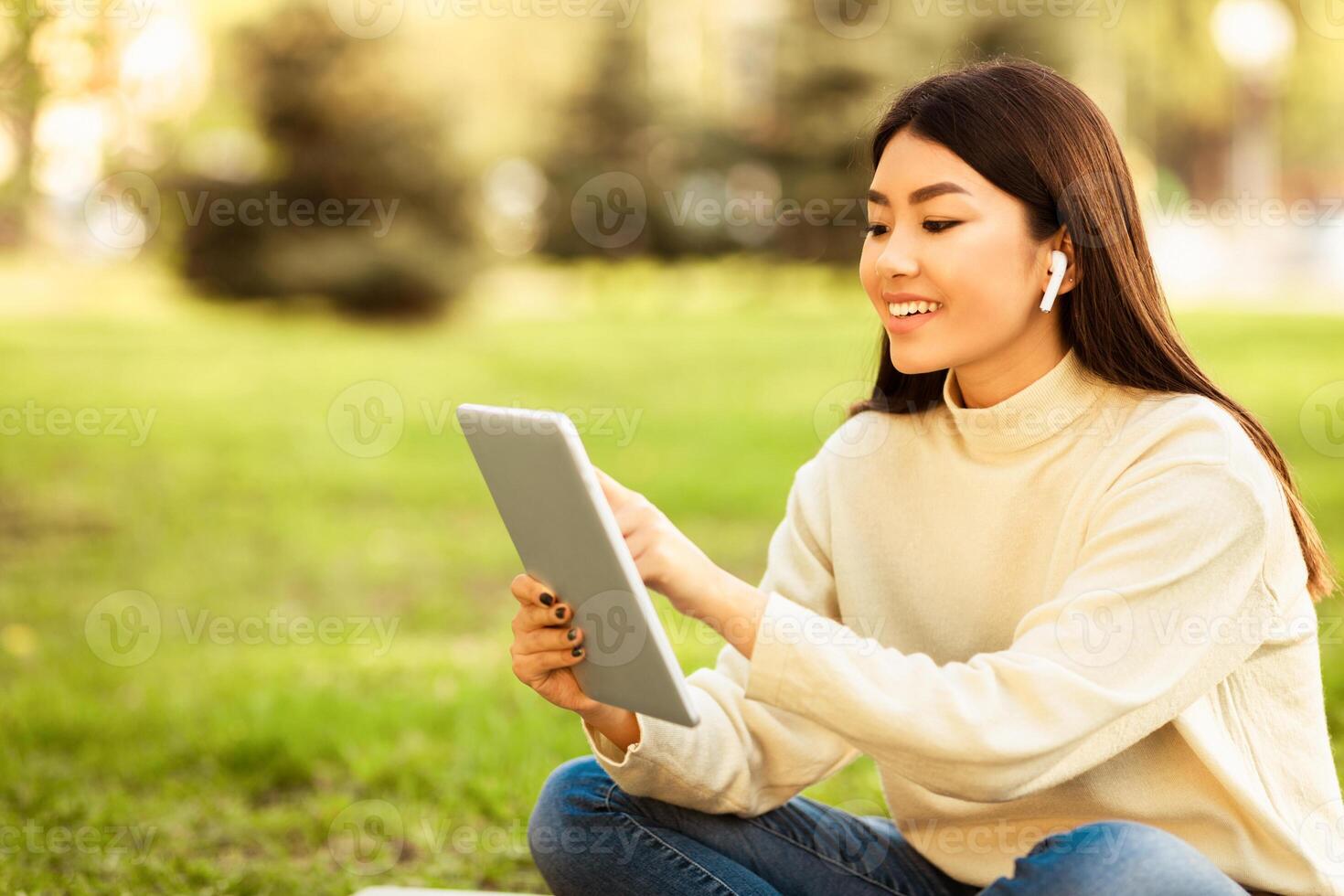 Asian woman sitting on the ground with legs crossed, focused on a tablet held in her hands. She appears engaged and concentrated as she interacts with the device, copy space photo