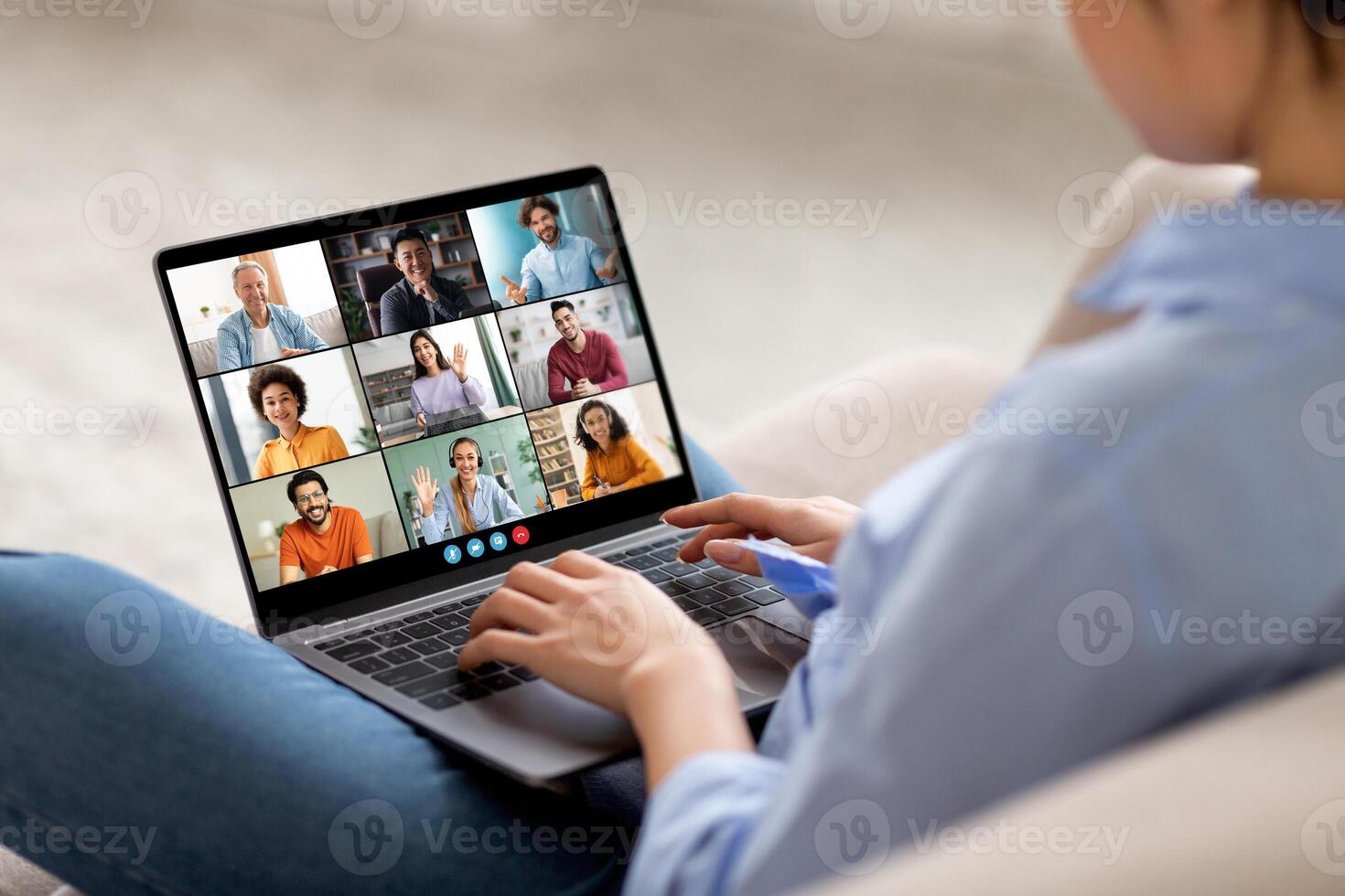 The image shows a woman sitting on a couch, participating in a conference on a laptop computer. Her laptop is open and shows a grid of nine feeds, each featuring a different person. photo