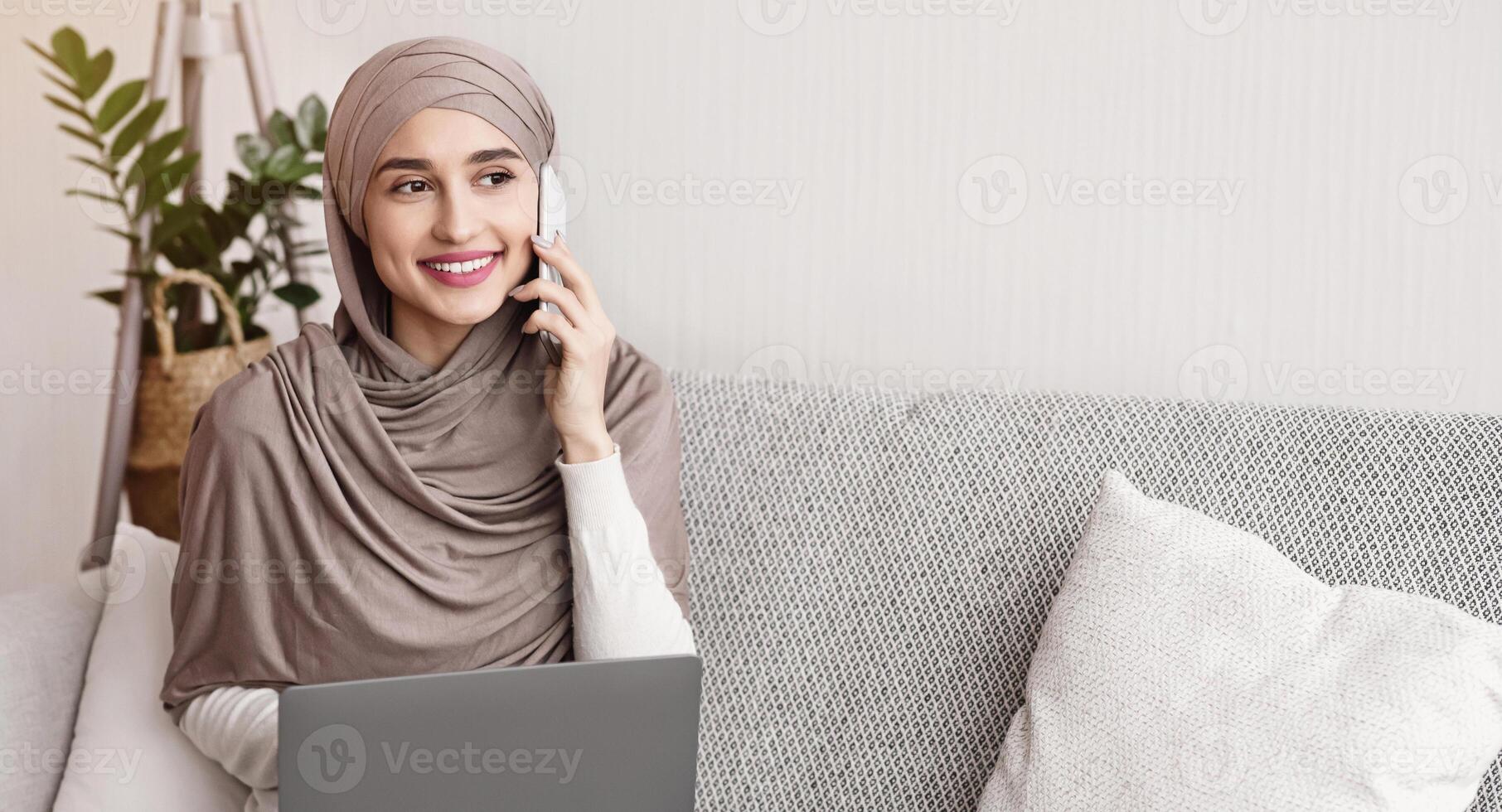 Arabic girl in headscarf sitting at home with laptop on lap and talking on cellphone, discussing details of her freelance work photo