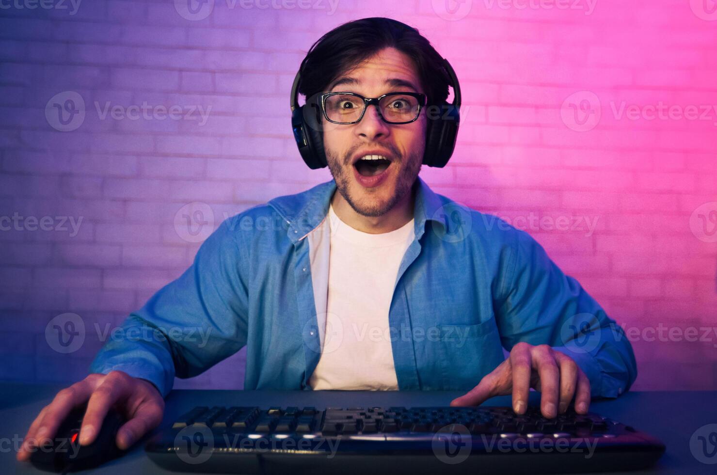 A young man wearing headphones and glasses is seated in front of a keyboard, looking surprised with his mouth open. A brick wall is behind him, and the room is lit with purple and pink lighting. photo