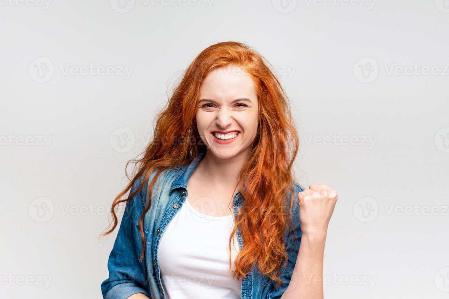 A young woman with vibrant red hair and a denim jacket is laughing and making a triumphant fist pump, symbolizing success or good news photo