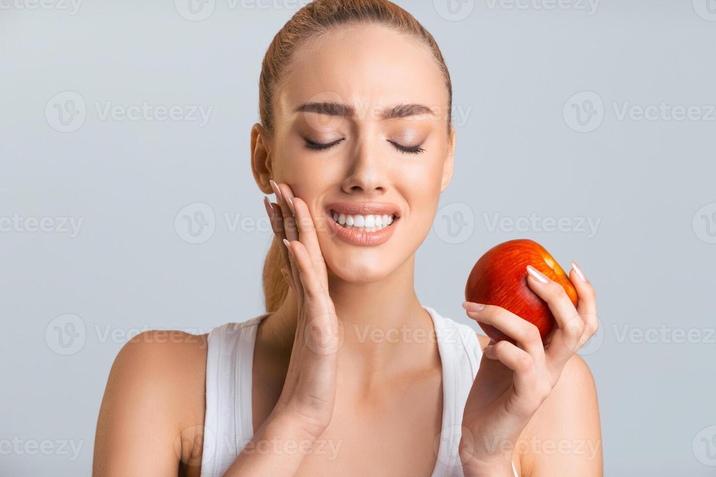 A woman with a white tank top is holding a red apple in her right hand. She is smiling and looking down at the apple. Her eyes are closed and her left hand is gently touching her cheek, have toothache photo