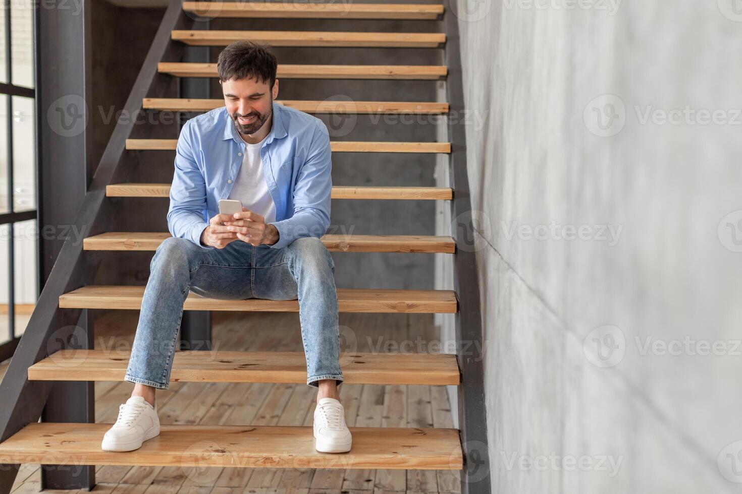 A man sits on a wooden staircase inside a building, smiling and looking at his smartphone. He is wearing blue jeans, a white t-shirt, and a blue button-down shirt. photo
