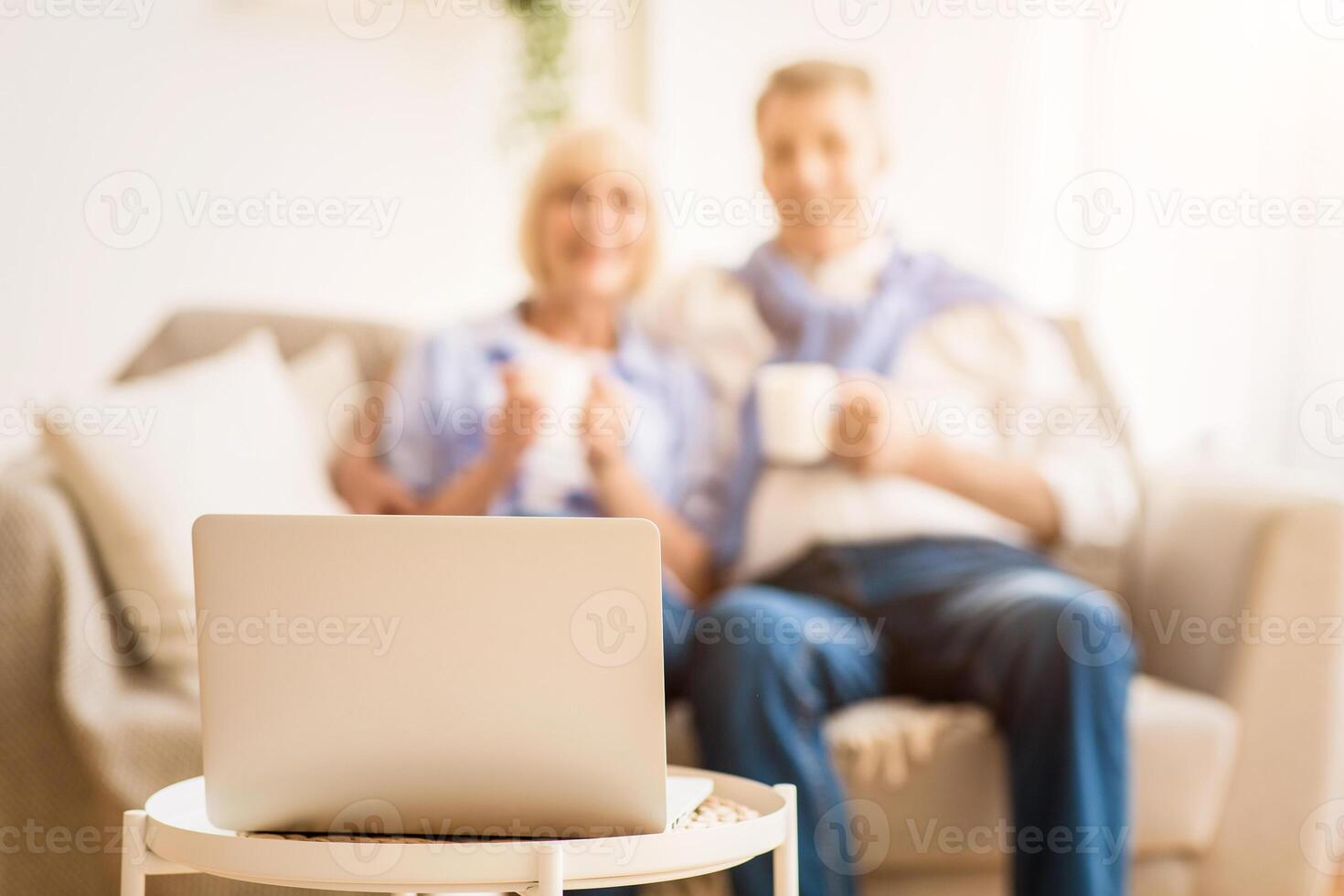 Senior couple watching tv online on laptop and drinking tea on comfortable sofa, focus on computer photo