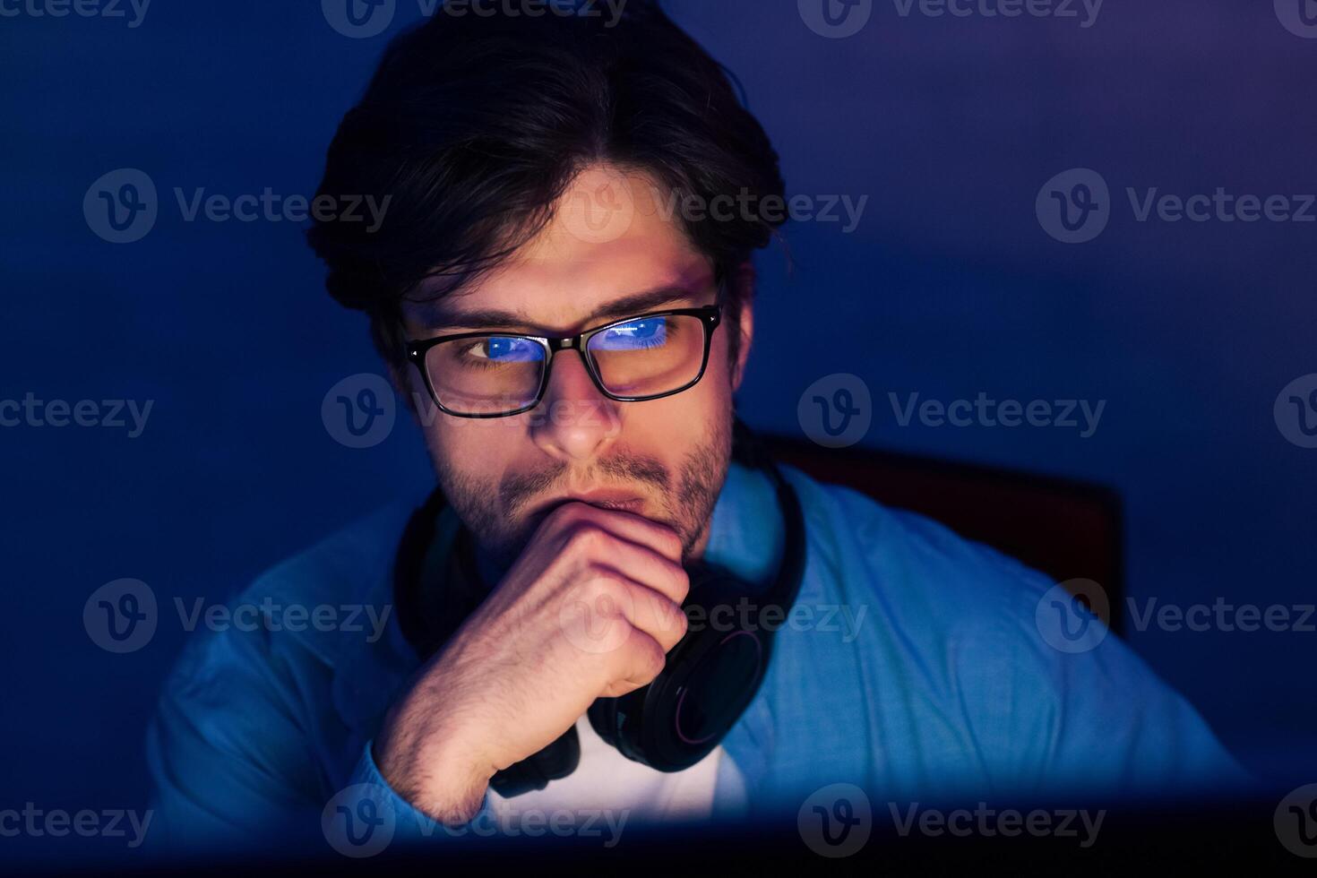A young man wearing glasses sits in front of a computer in a dark room, thoughtfully resting his chin on his hand, likely deep in concentration on a project or task. photo