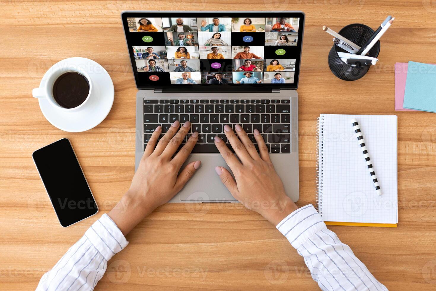 A person is sitting at a wooden desk and using a laptop to participate in a conference. The laptops screen shows a grid of tiles, each displaying a different participant, top view photo