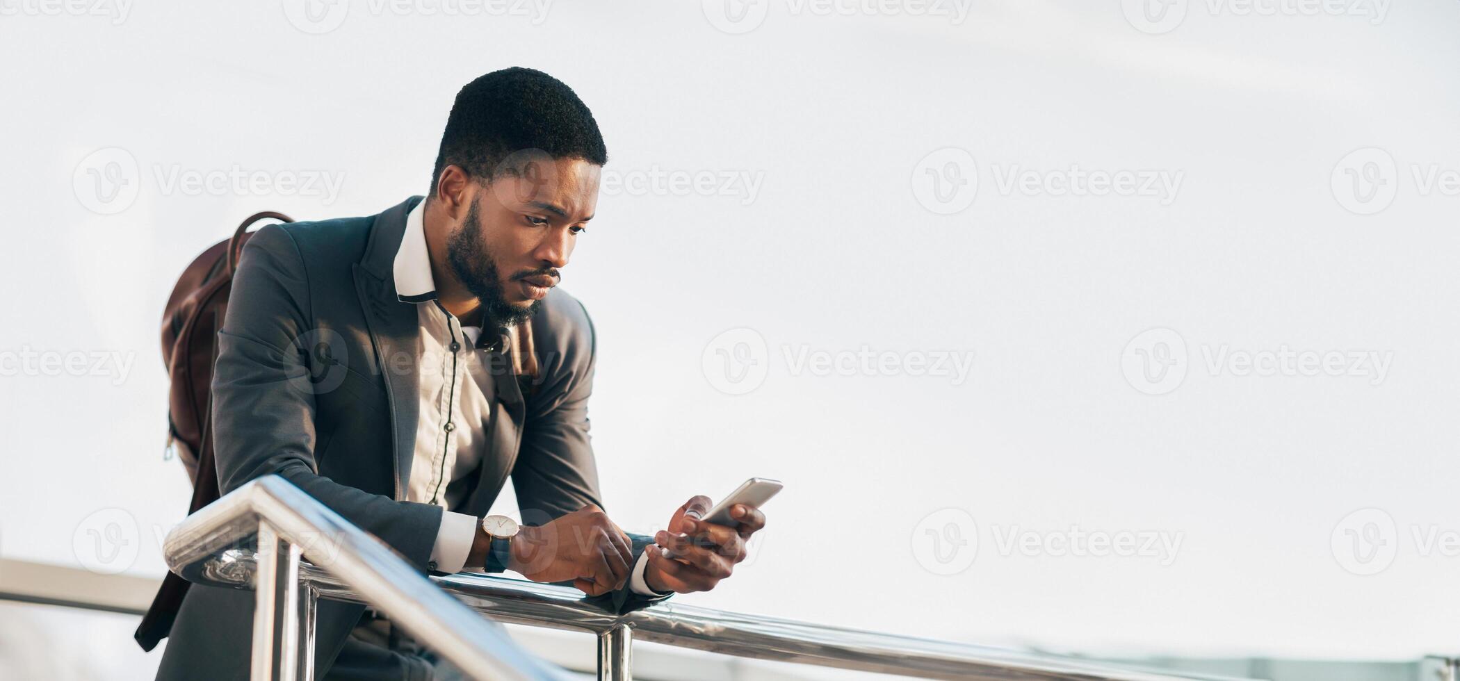 Black guy entrepreneur in a suit checking his phone while standing with a rolling suitcase outside a modern building, panorama with copy space photo