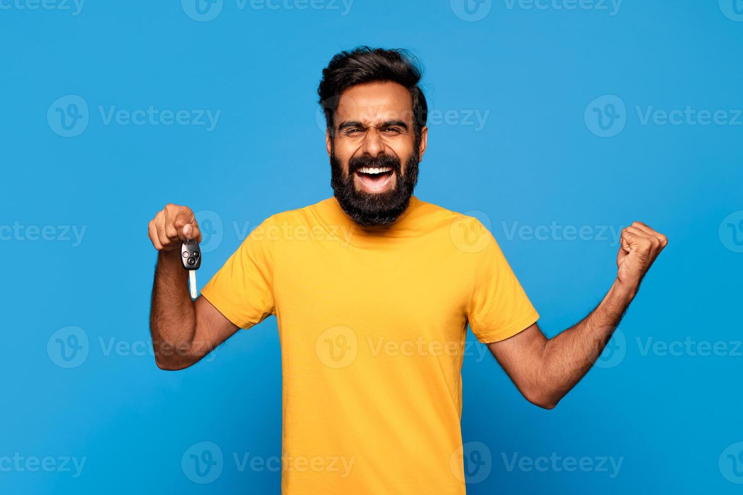 Hindu man with a beard is smiling and holding up a car key in his right hand while his left fist is clenched in the air. He is wearing a yellow t-shirt and is standing in front of a blue background. photo