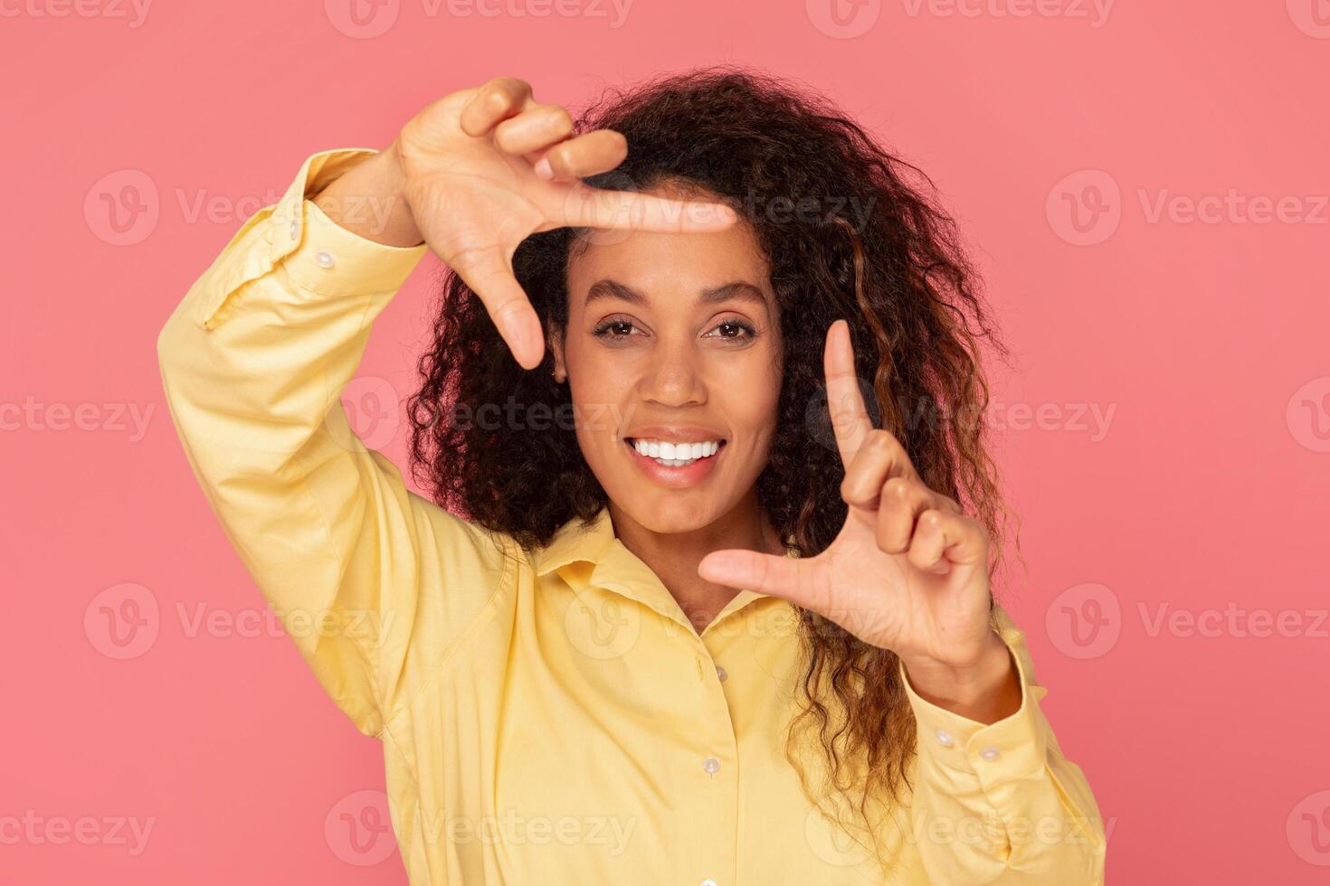 A young black woman with curly hair is smiling at the camera while making a framing gesture with her hands in front of her face, the background is a solid pink. photo