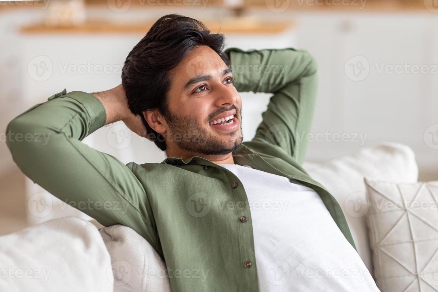 Arab man with a beard is sitting on a white couch with his arms behind his head, looking relaxed and content. The background appears to be a living room. photo