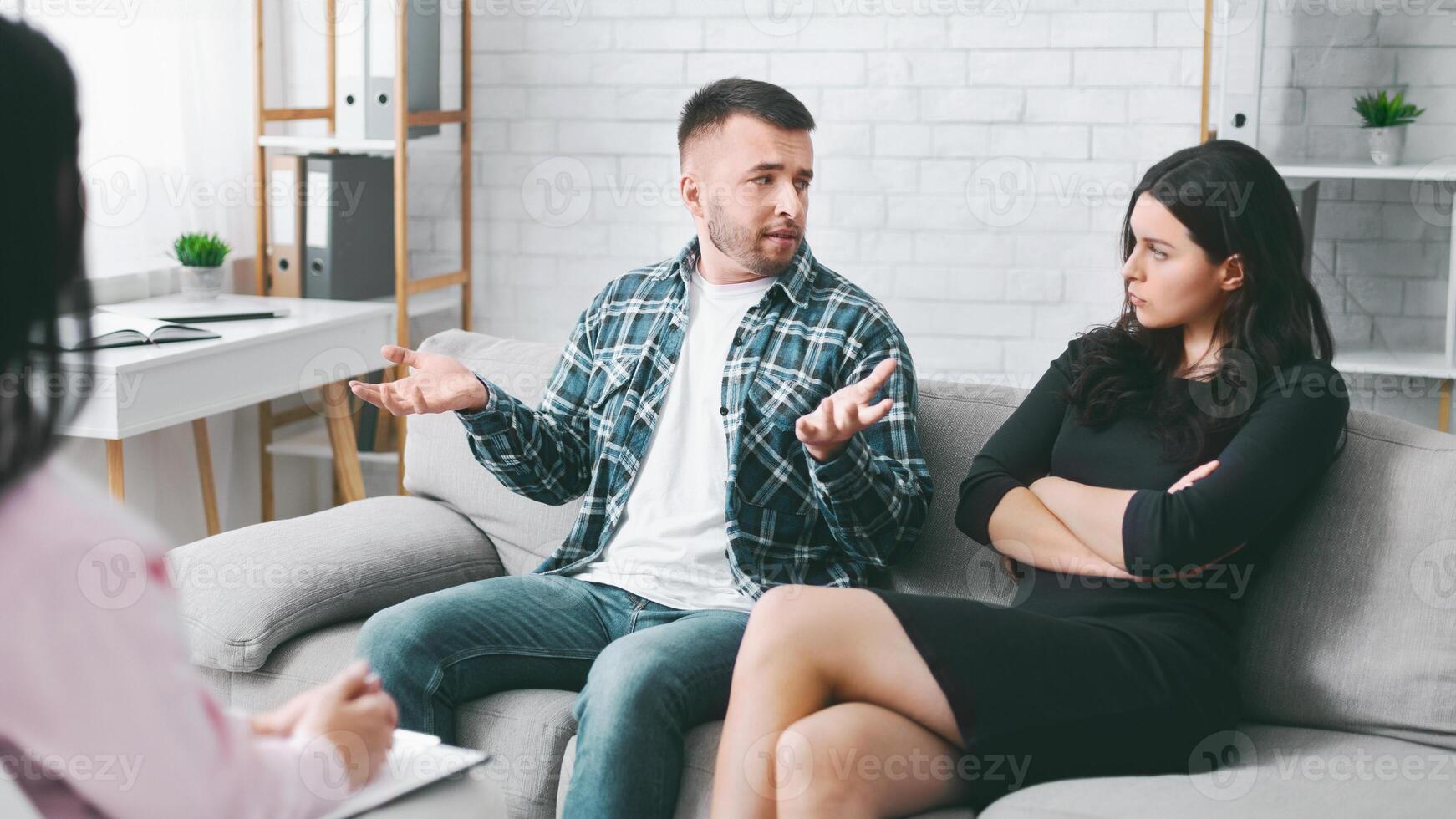 This image shows a couple in the middle of a heated argument during a therapy session. The man is gesturing wildly with his hands, while the woman has her arms crossed and looks at him photo