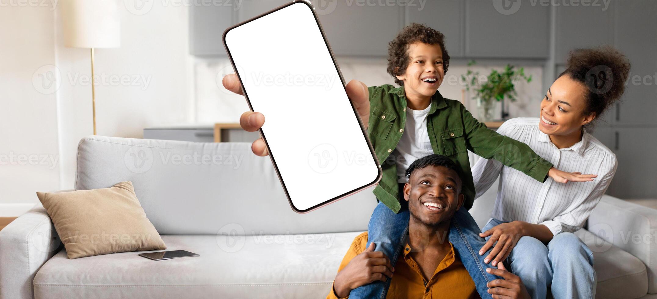 A joyful African American family interacts at home, with a child playfully seated on a parent shoulders, while another parent holds up a smartphone, capturing a happy moment together. photo
