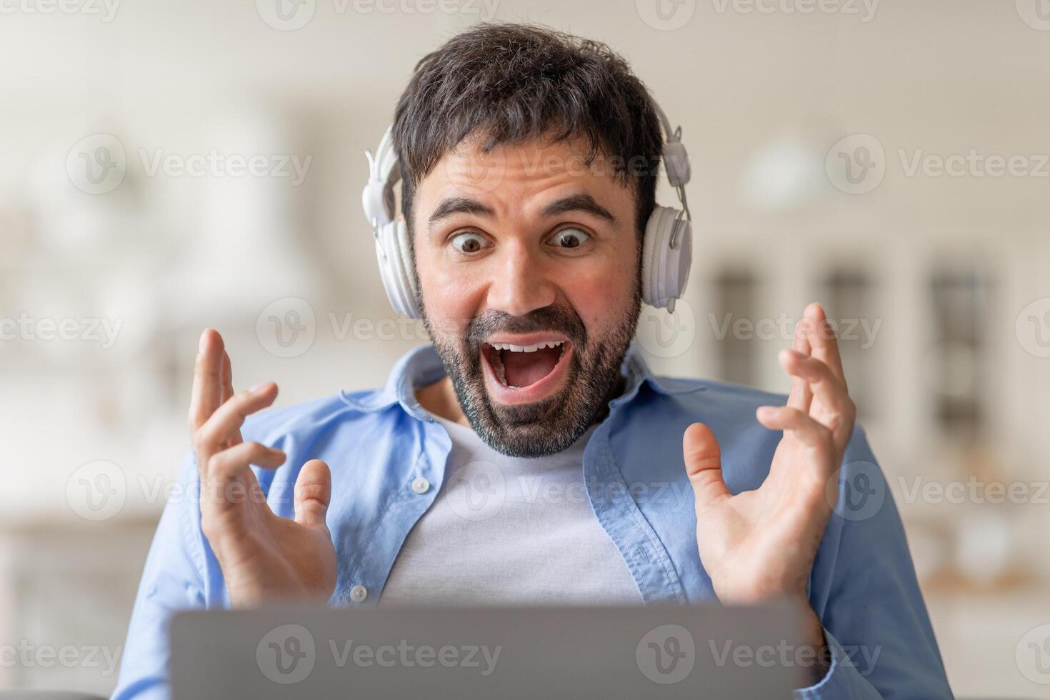 A man wearing headphones sits in front of a laptop with a wide open mouth and his hands raised in the air, showing excitement. He is looking at screen and appears to be engaged in something exciting. photo