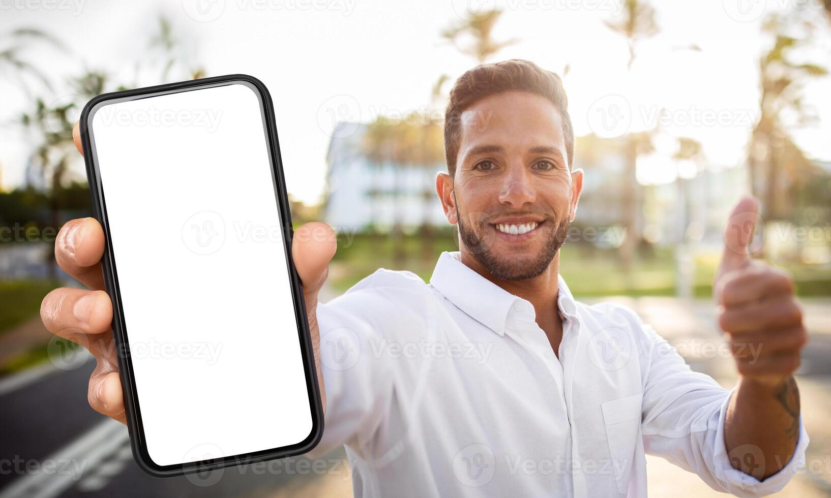 A man is standing outdoors, holding a smartphone with a blank screen, and giving a thumbs-up. He is smiling and looking directly at the camera. photo