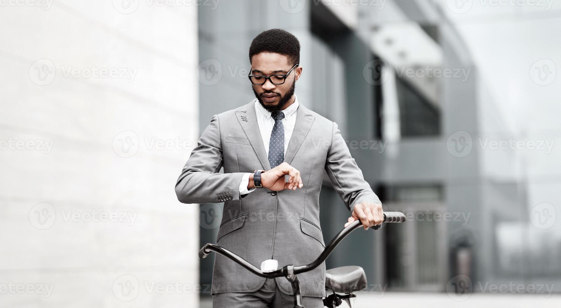 Black businessman in a suit on a bicycle checking his watch in an urban setting, going to business meeting, copy space photo