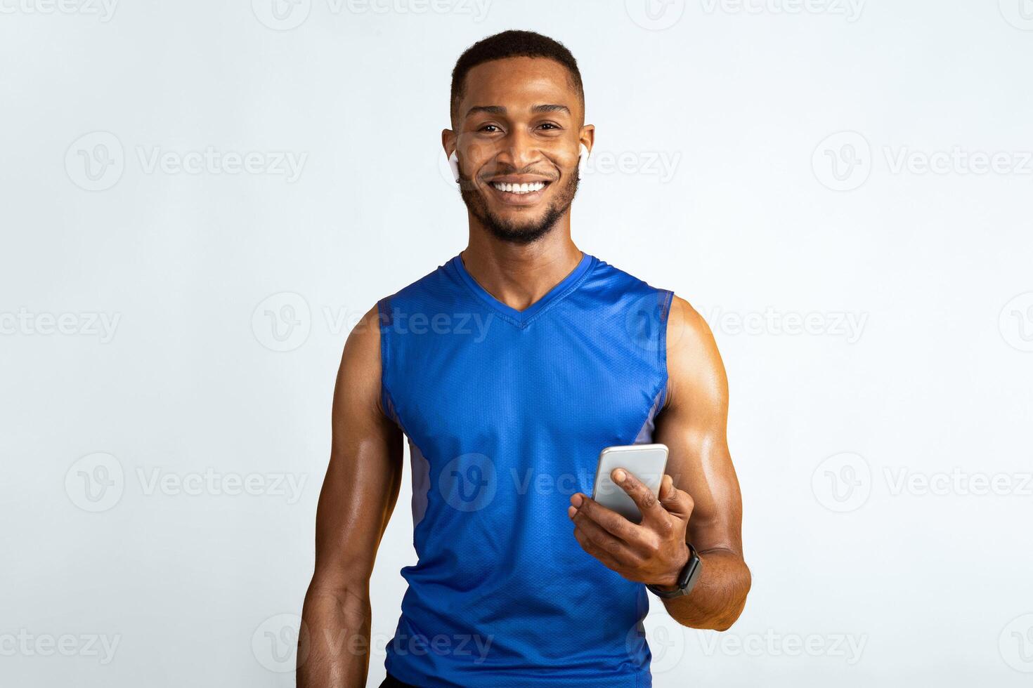 Working Out. Smiling afro man in earbuds holding smartphone, using fitness app isolated on yellow studio wall, copyspace photo