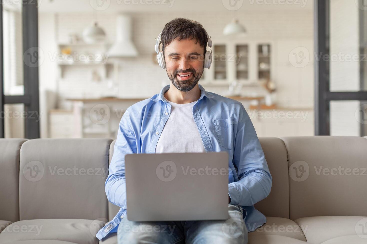 A man sits on a light gray couch in a modern living room, smiling while working on his laptop. He is wearing headphones and a light blue button-up shirt. photo