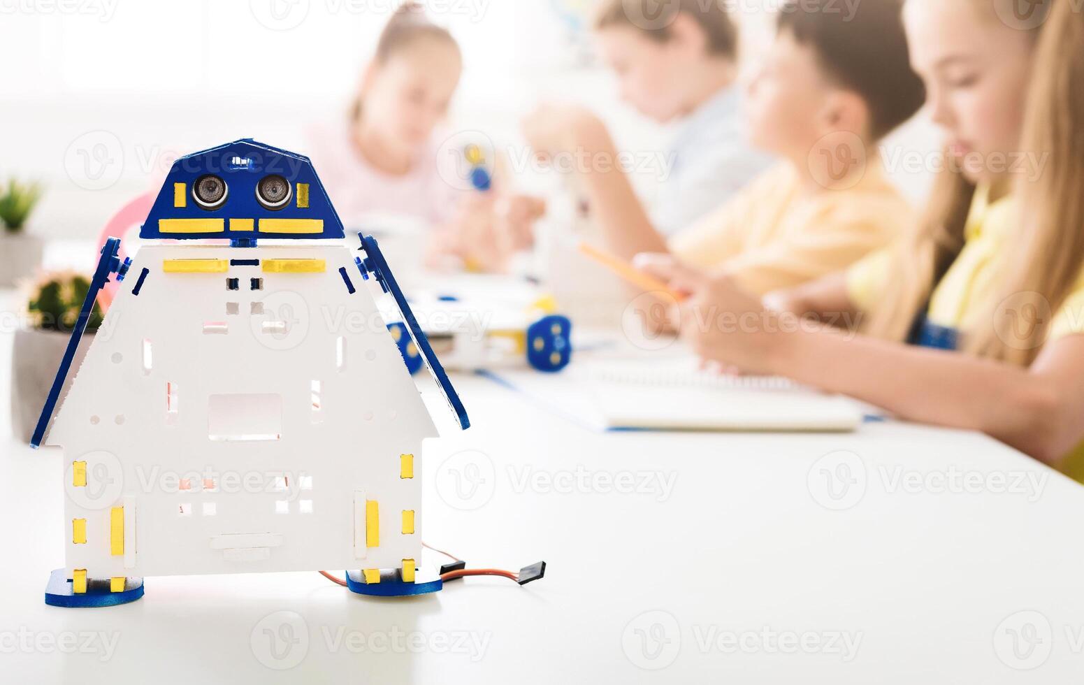A close-up shot of a white and blue robot toy sitting on a table in front of a classroom of children. The children are out of focus in the background, and the robot toy is in sharp focus. photo