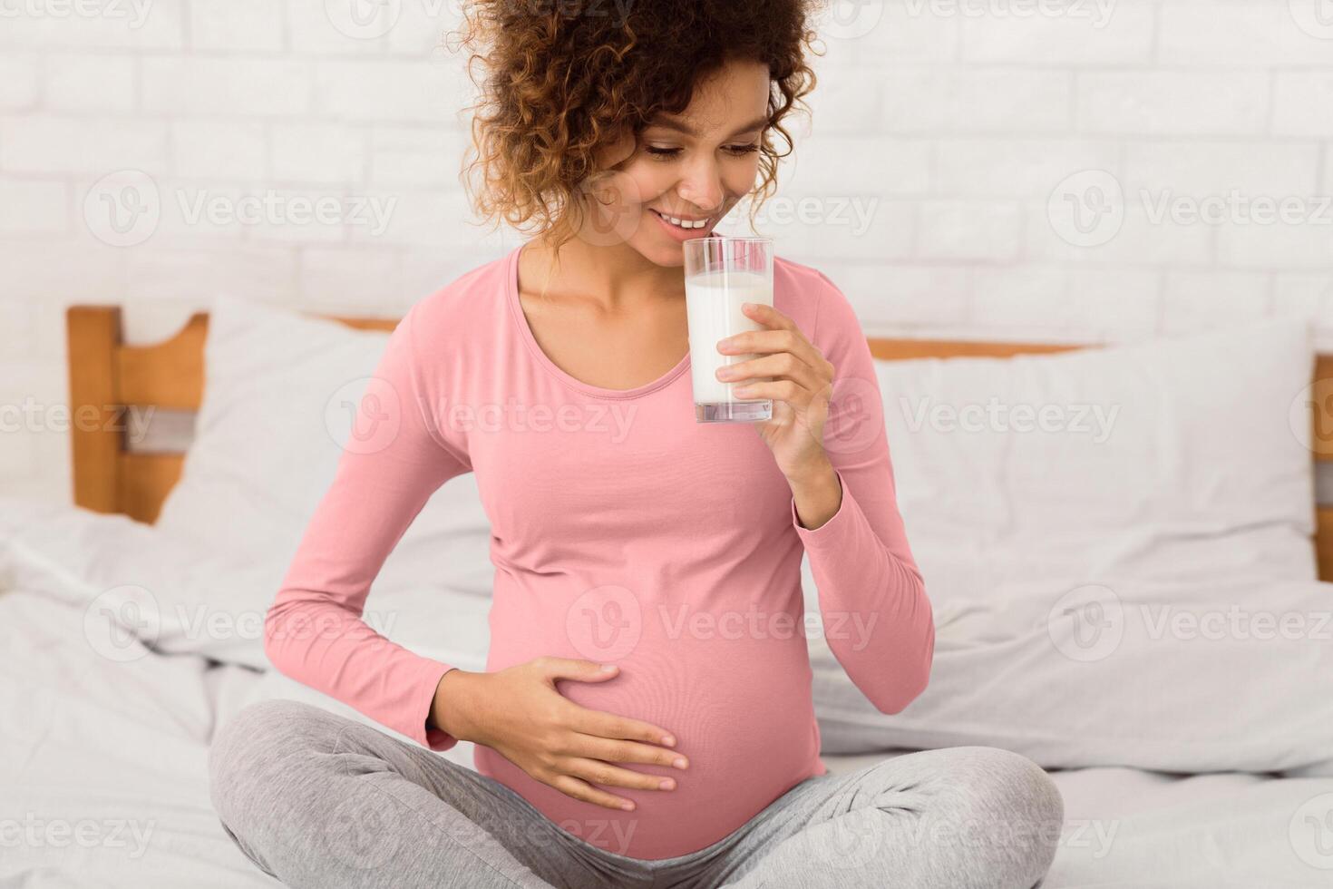 Useful diet for pregnant. Afro woman with glass of milk, sitting on bed in the morning photo