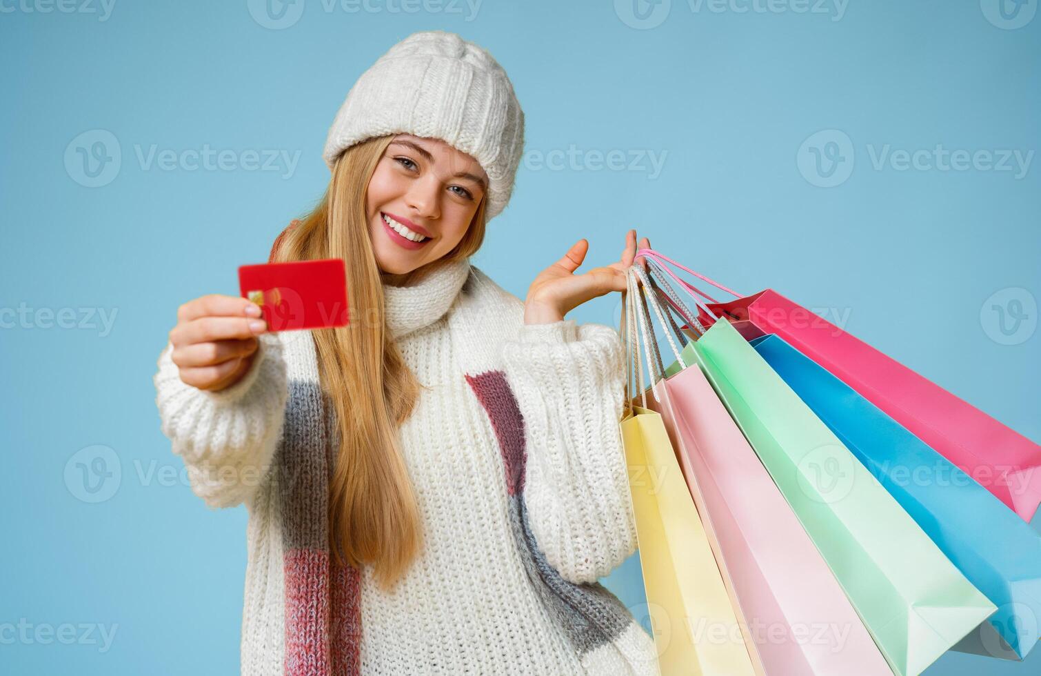 Happy young woman in white hat recommending credit card, posing with shopping bags and looking at camera, yellow background, free space photo