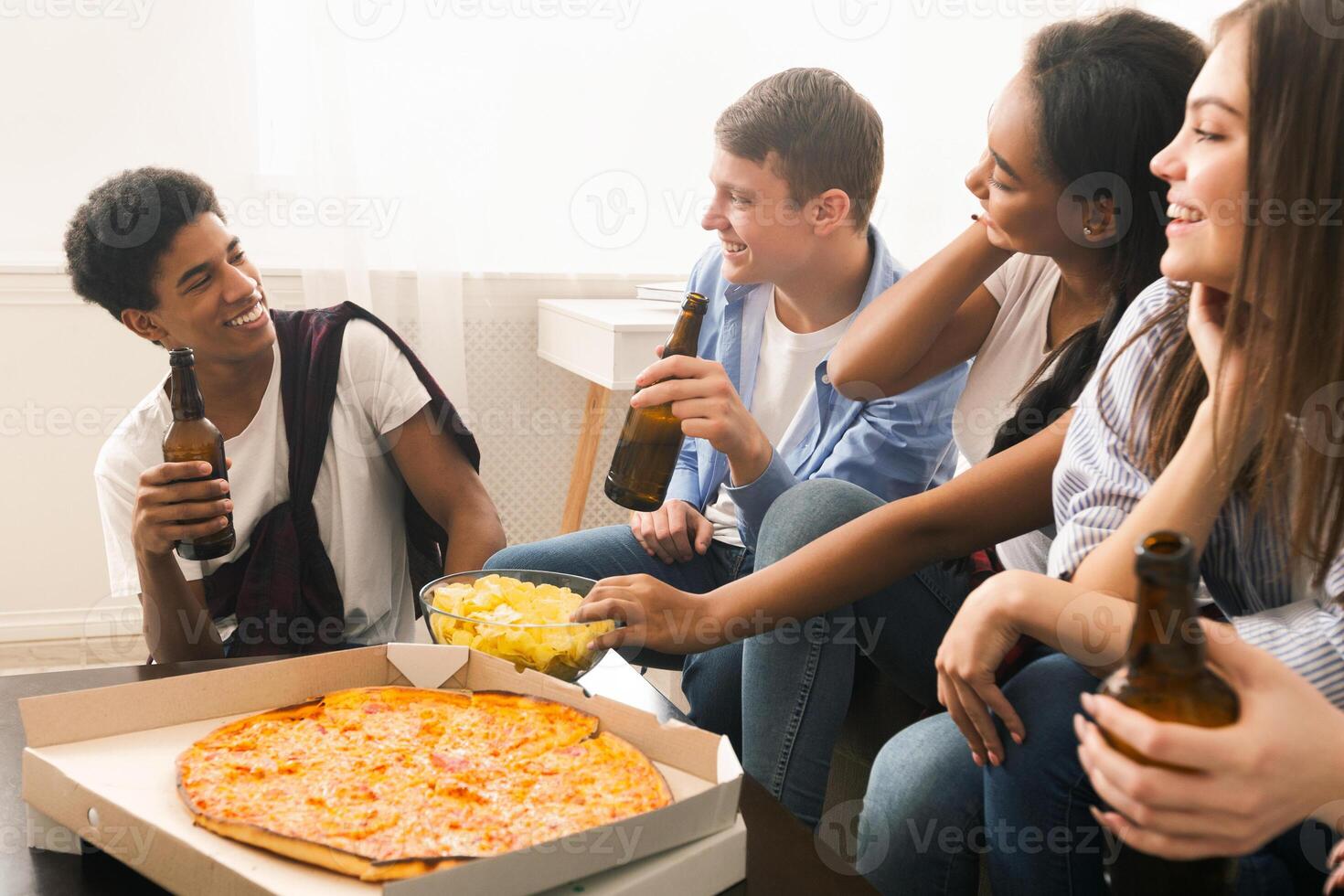 A group of diverse friends sitting in a circle around a large box of pizza, engaged in conversation and reaching out to grab a slice. The pizza box is open, revealing a variety of toppings. photo