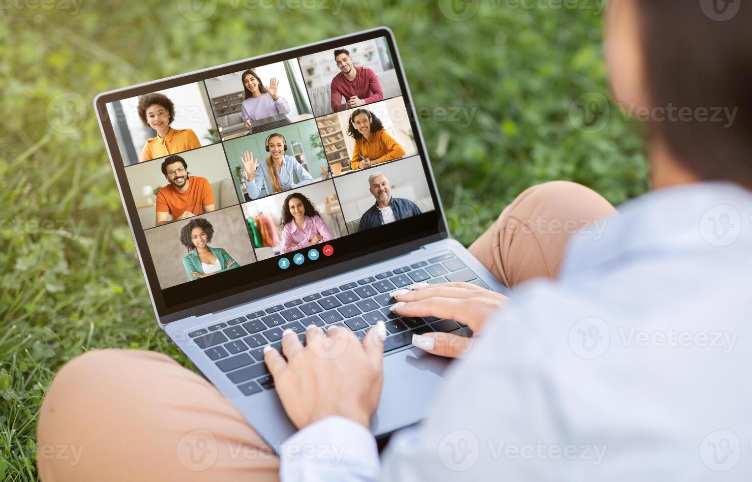 A person sits outside on a grassy lawn, using a laptop for a conference. The screen displays a grid of nine people, each in a separate frame photo