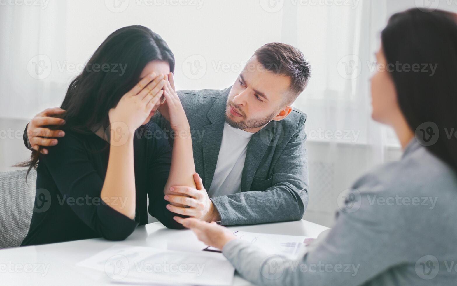 The image shows a man comforting a woman who is upset and covering her face with her hands. They are sitting at a table with a counselor, likely discussing a difficult situation. photo