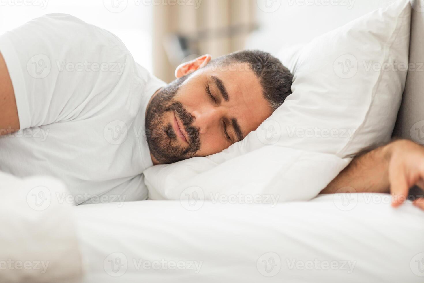 A man with a beard sleeps peacefully on his side, his head resting on a white pillow and his arm outstretched. The bedding is all white and the room appears to be a bedroom. photo