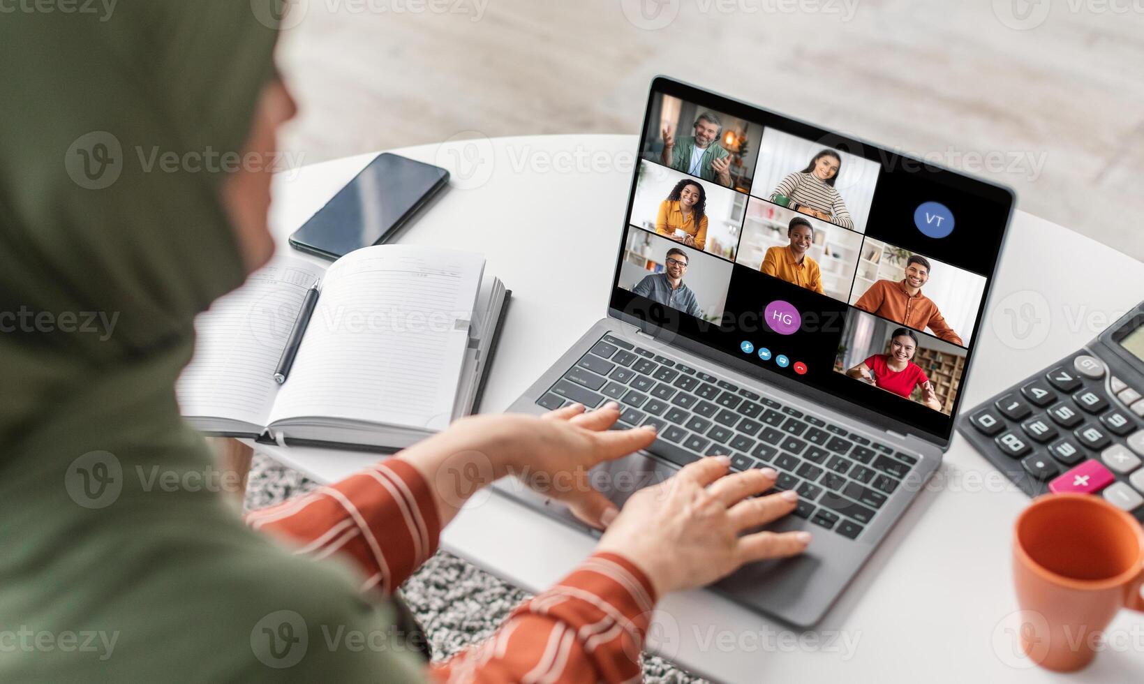 A woman in a green hijab is sitting at a desk with a laptop. She is looking at the screen which is showing a conference call with multiple people. photo