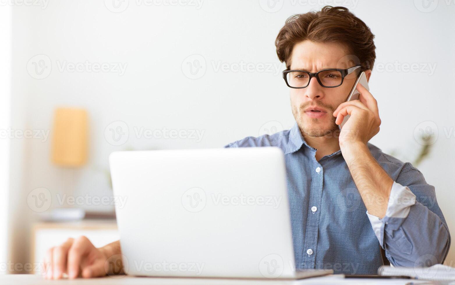 A man in a blue shirt and glasses sits at a desk, talking on his phone while looking at his laptop. photo